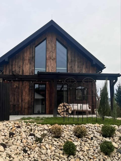 Nowoczesny dom z drewna o ciemnej elewacji, z dużymi oknami i ozdobnym ogrodem skalnym na pierwszym planie. Widoczne drewno i elementy dekoracyjne.