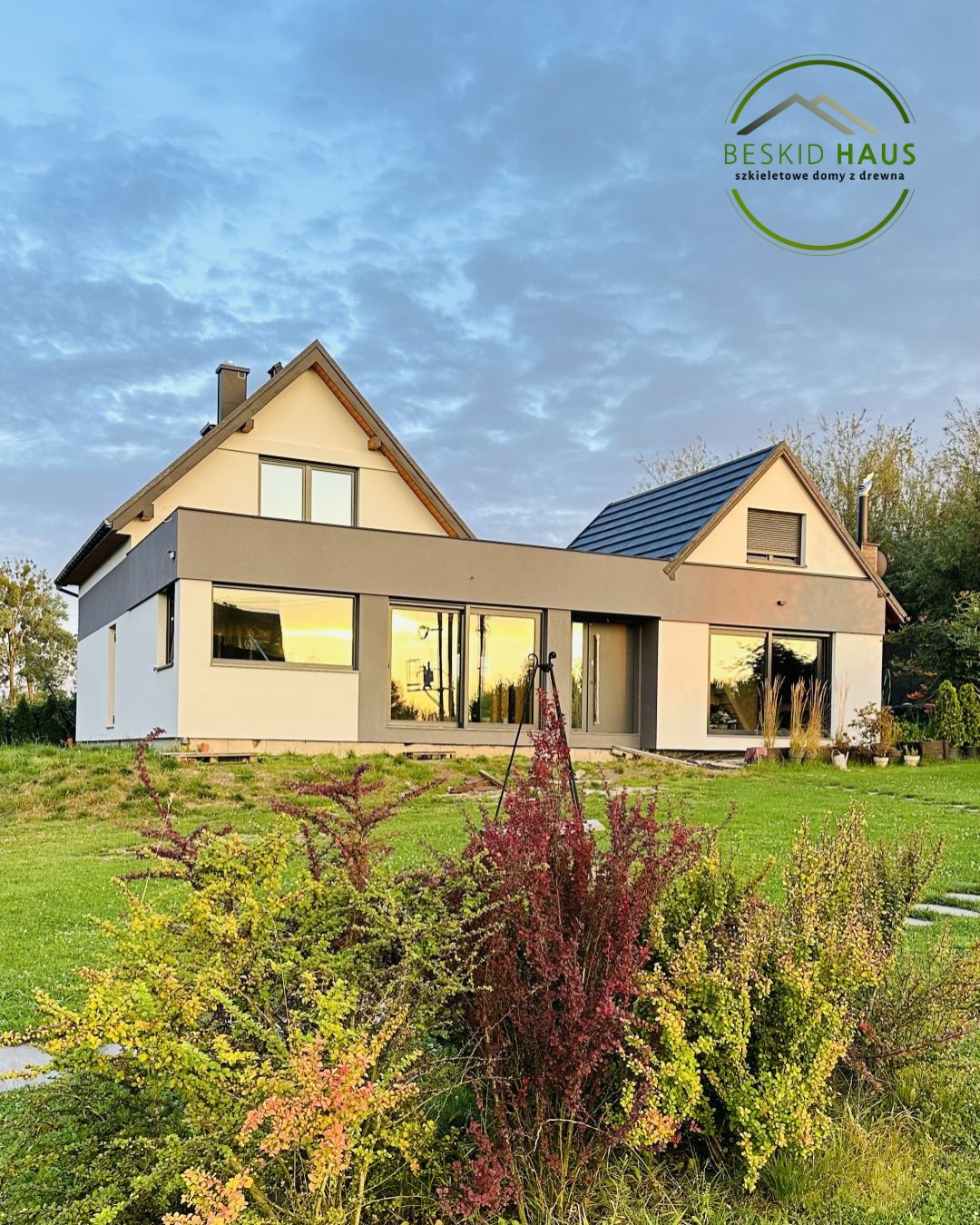 Nowoczesny dom z elementami szkieletowymi, z dużymi oknami odbijającymi zachód słońca, otoczony zielenią i ozdobnymi krzewami. Widok z poziomu ogrodu.