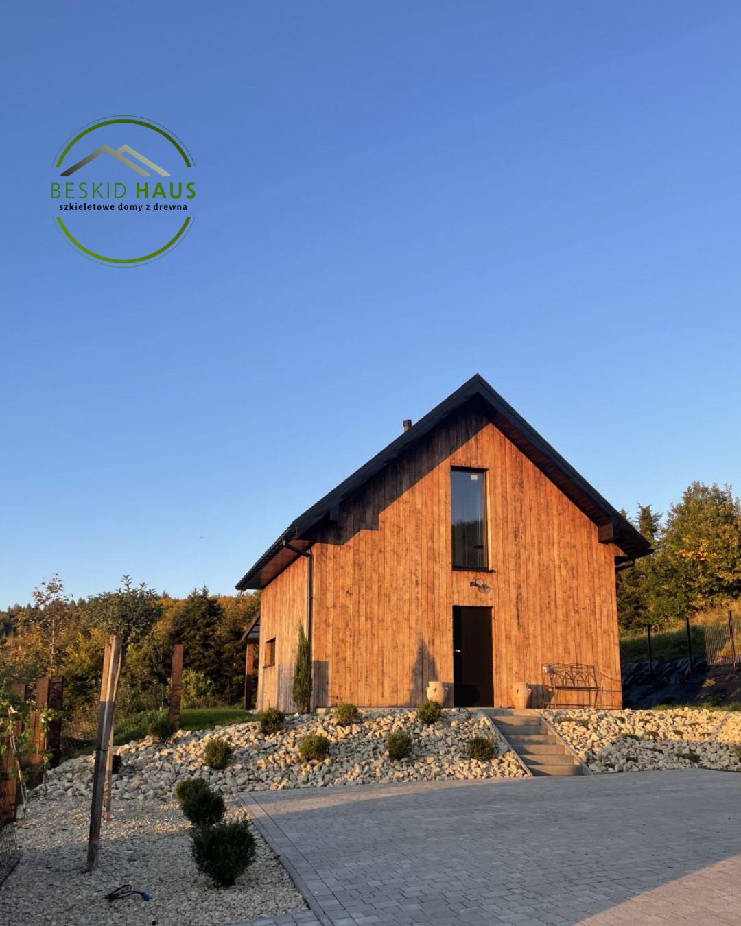 Nowoczesny dom z drewna o prostej bryle, z dużym oknem nad wejściem, otoczony kamieniami i zielenią, w ciepłym świetle zachodzącego słońca.