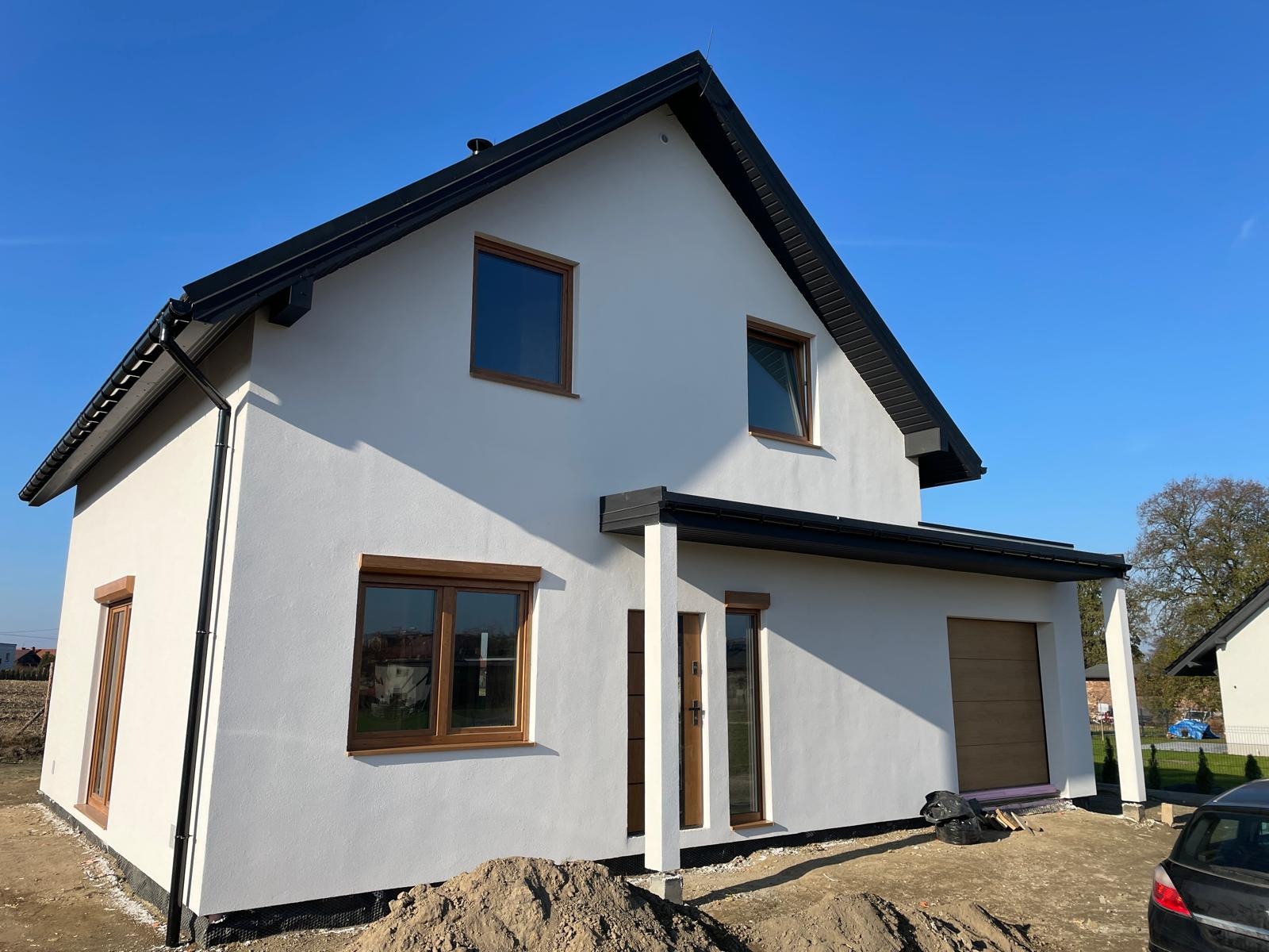 Biały dom z czarnym dachem i brązowymi oknami w stylu nowoczesnej stodoły, z widocznym fragmentem samochodu i stertą ziemi przed budynkiem.