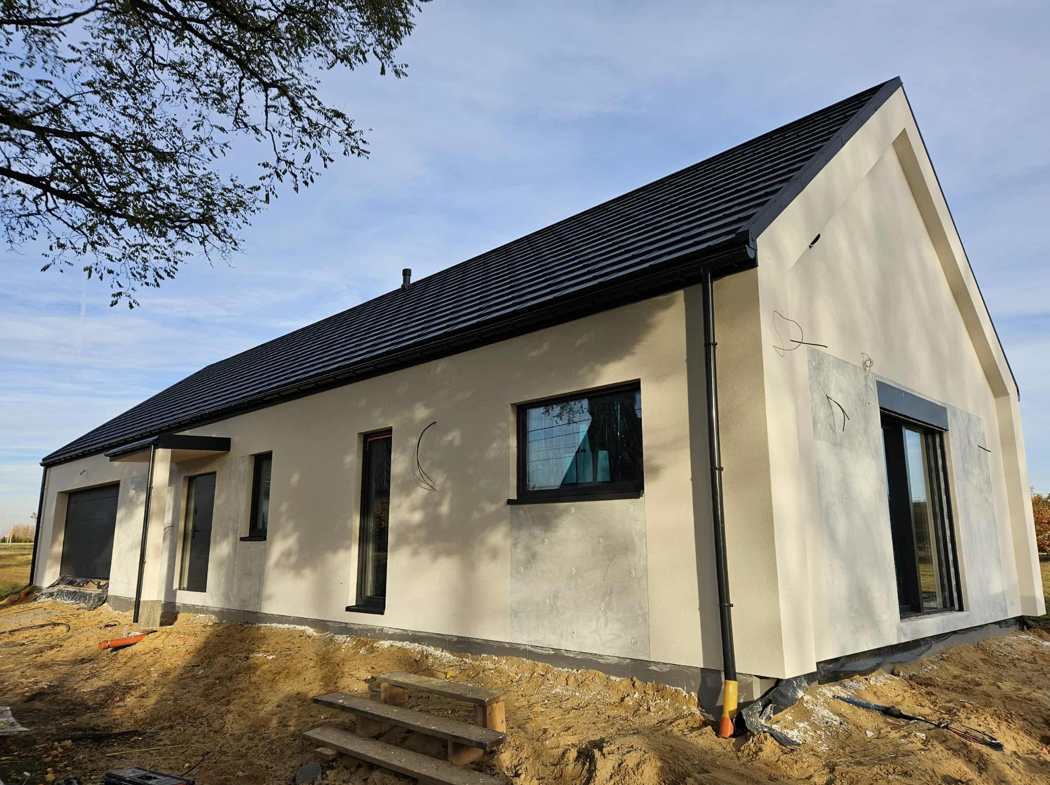 Nowoczesny dom jednorodzinny z czarnym dachem i oknami, w trakcie wykończenia elewacji. Widoczne przygotowania terenu wokół budynku i fragment drzewa w tle.
