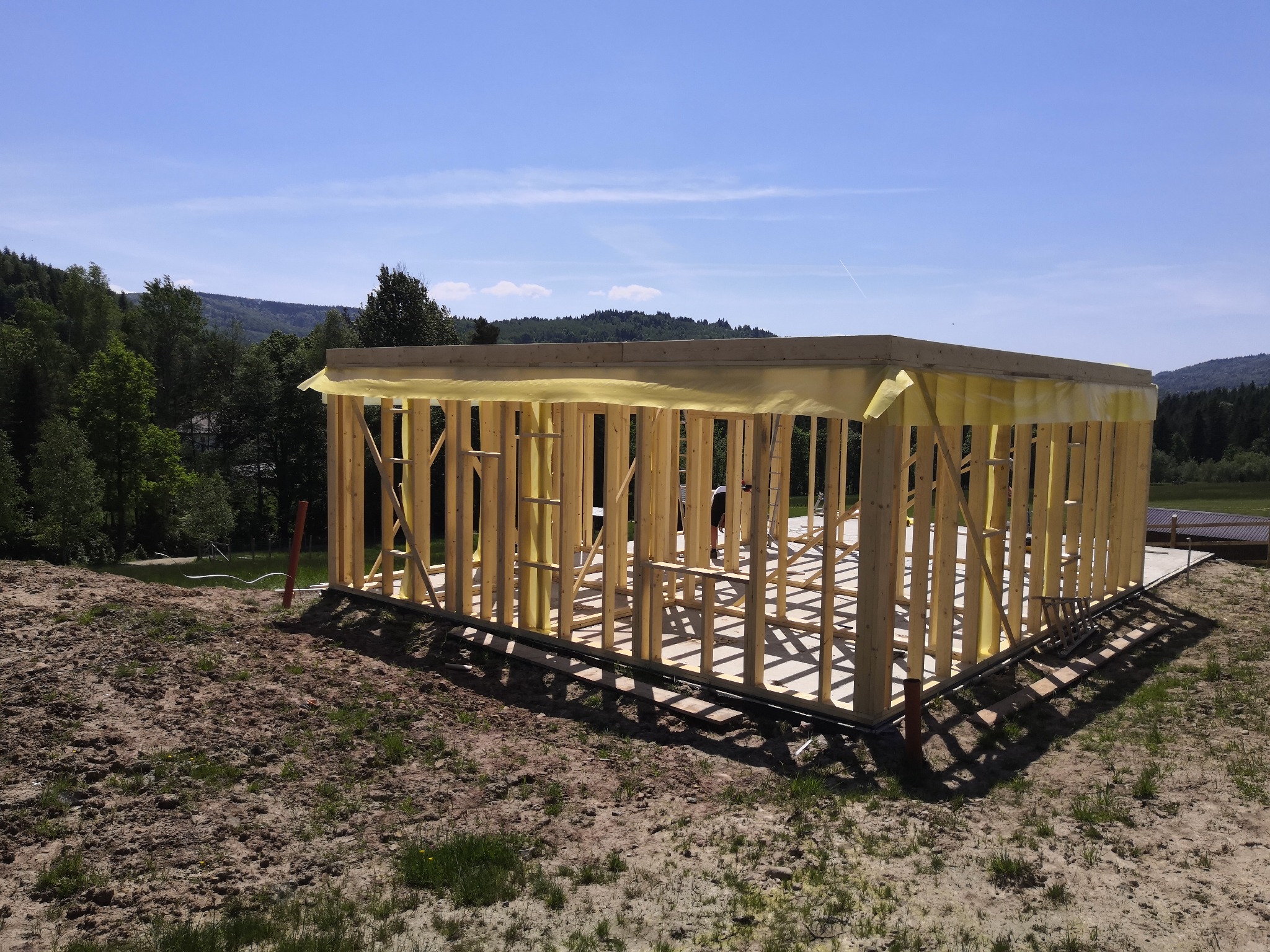 Surowa konstrukcja domu szkieletowego z widoczną żółtą folią wiatroizolacyjną, na tle zalesionych wzgórz i błękitnego nieba.