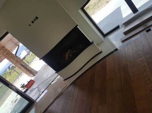 Ukończona podłoga z ciemnego drewna w salonie z kominkiem, widoczne listwy przypodłogowe i fragment okna z widokiem na zewnątrz.