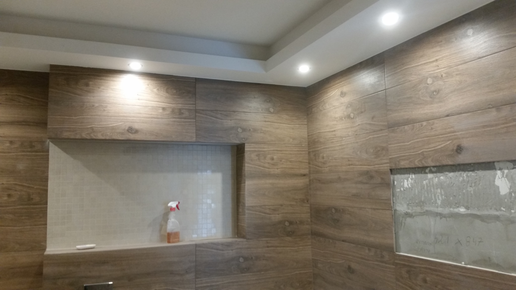 Łazienka w trakcie wykańczania, ściany wyłożone płytkami imitującymi drewno, wnęka z mozaiką, widoczna butelka ze środkiem czyszczącym, obok fragment ściany bez płytek z widocznym betonem...
