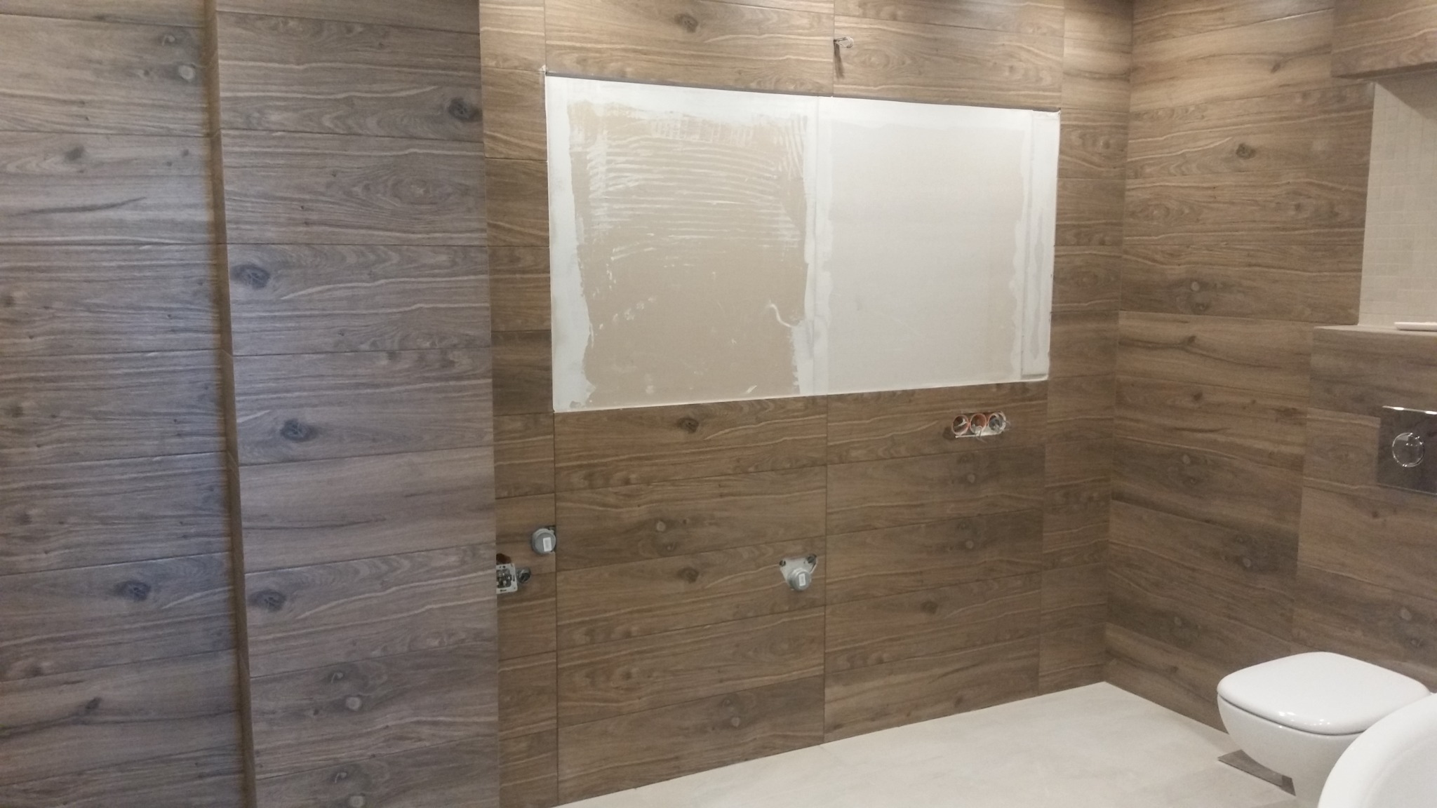 Łazienka w trakcie remontu: drewnopodobne płytki ścienne, częściowo położone, z widocznymi otworami na armaturę i fragmentem ściany przygotowanej pod glazurę.