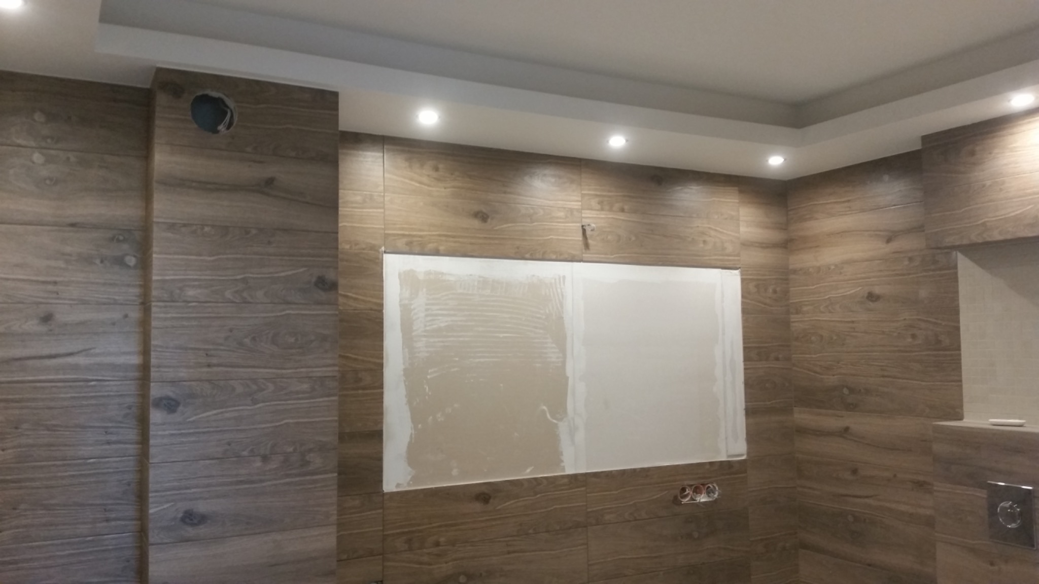 Ściana wyłożona panelami imitującymi drewno z fragmentem przygotowanym pod płytki, widoczne gniazdka elektryczne i oświetlenie sufitowe