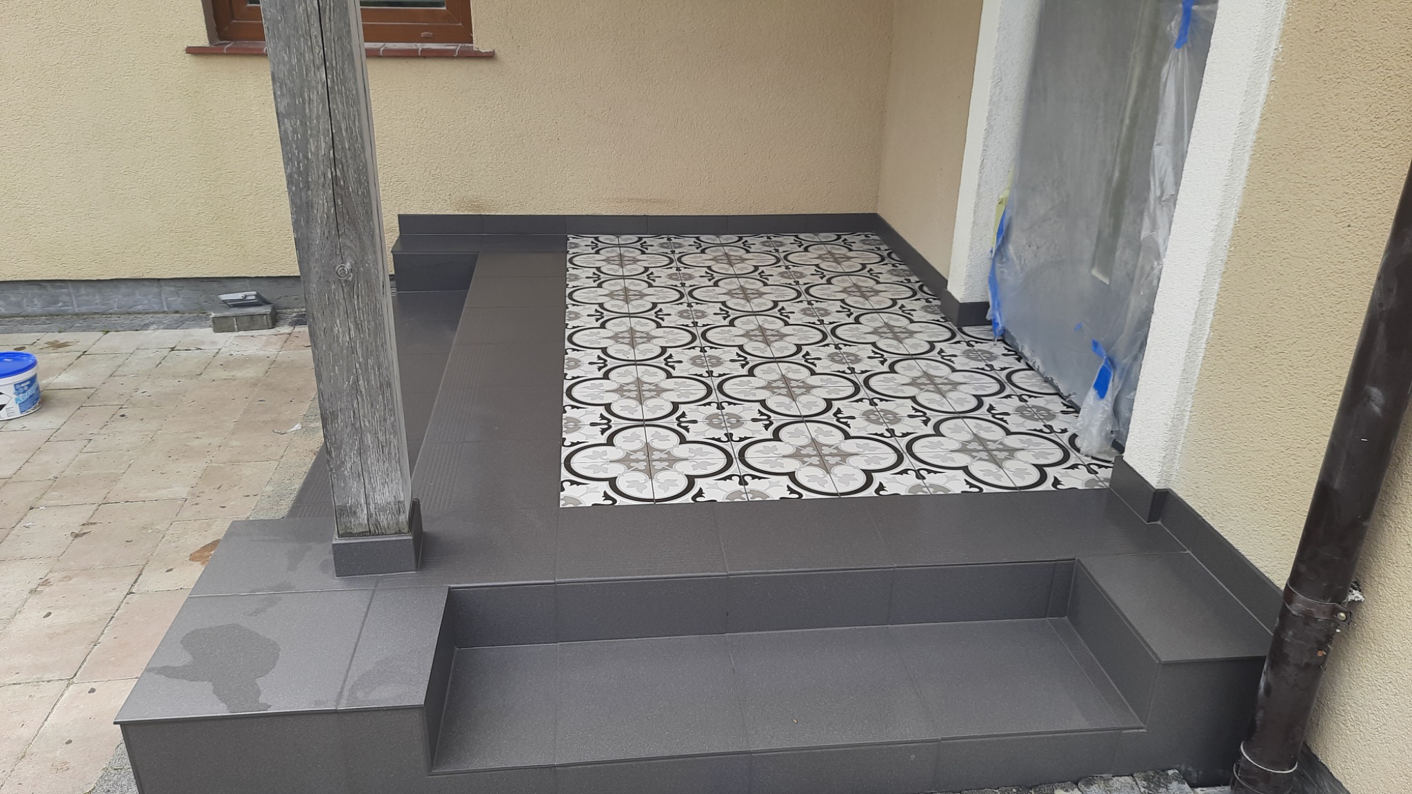 Wykończone schody wejściowe z ciemnoszarych płytek z dwoma stopniami, wyłożone płytkami w geometryczny wzór, z drewnianą podporą z boku i zabezpieczonym wejściem folią malarską.