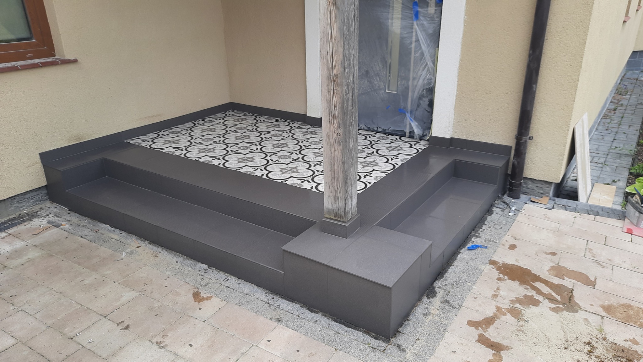 Wykończone schody wejściowe do domu z szarych płytek ceramicznych, z ozdobną posadzką na podeście i drewnianym słupem podtrzymującym zadaszenie, drzwi wejściowe zabezpieczone folią.