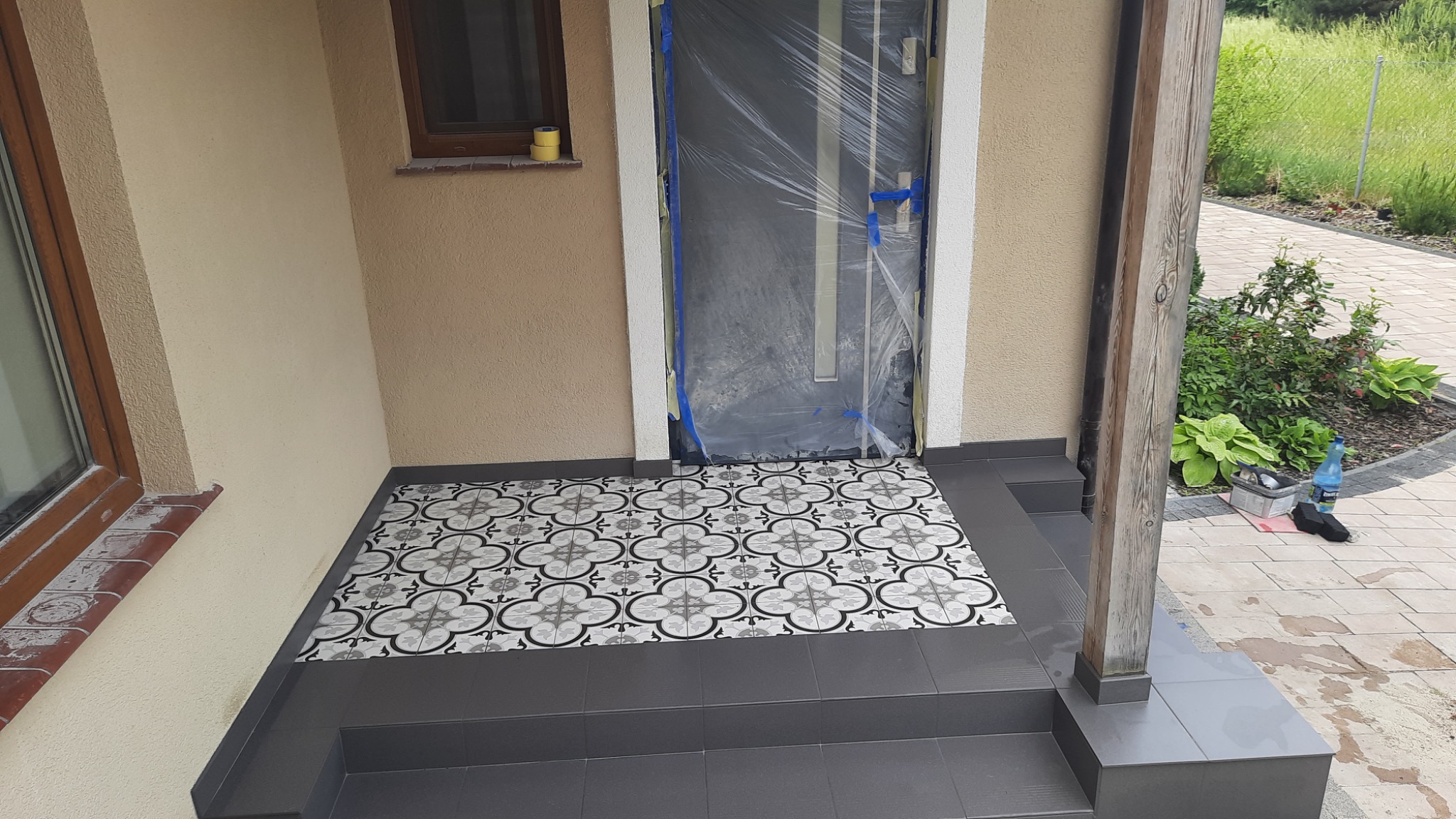 Wyłożona wzorzystymi płytkami posadzka na schodach wejściowych, zabezpieczone folią drzwi, jasna elewacja budynku, widoczne narzędzia i materiały budowlane.