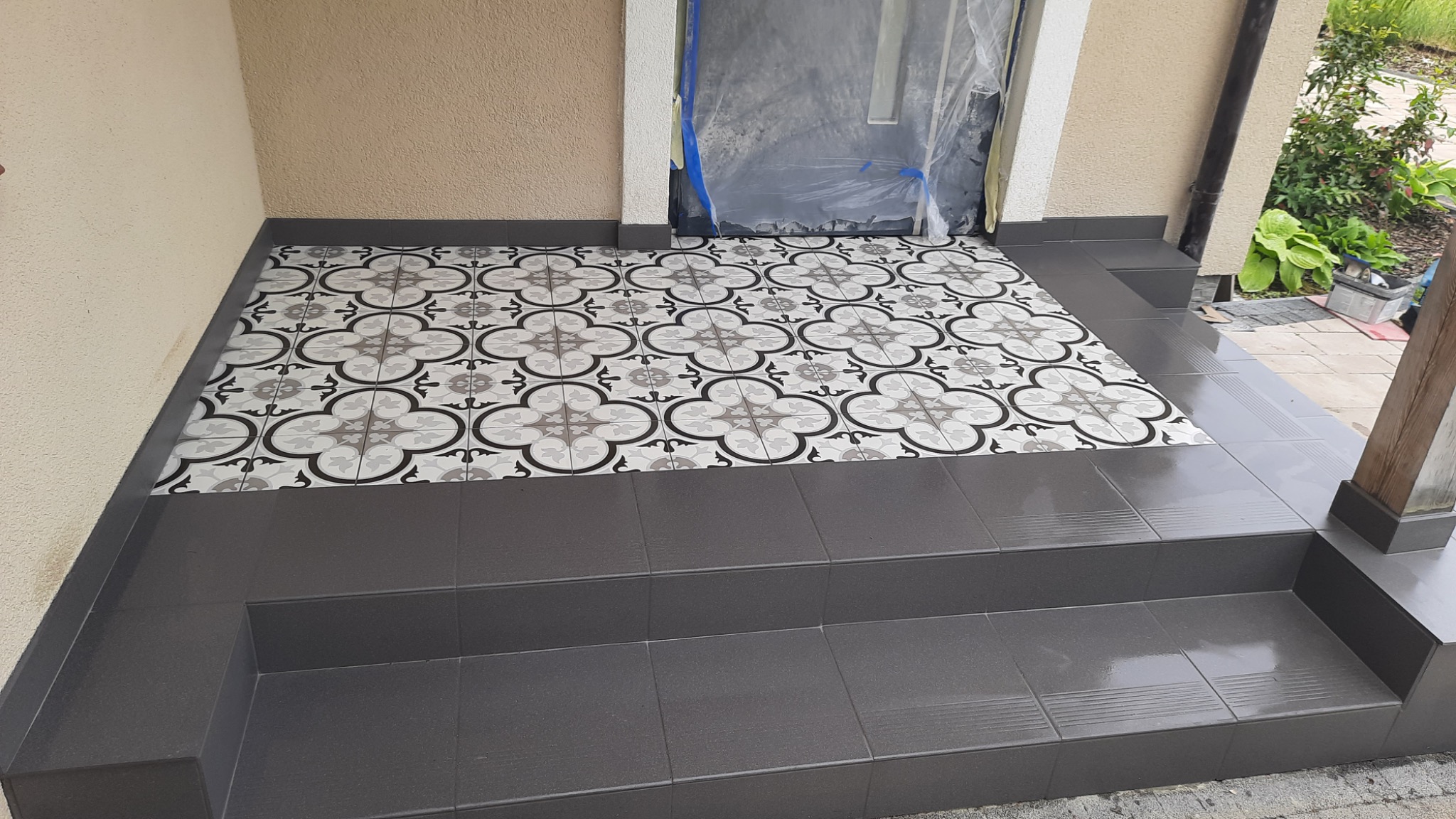 Wykończone płytkami w geometryczny wzór schody wejściowe do domu, z zabezpieczonym folią wejściem i widoczną zielenią w tle.