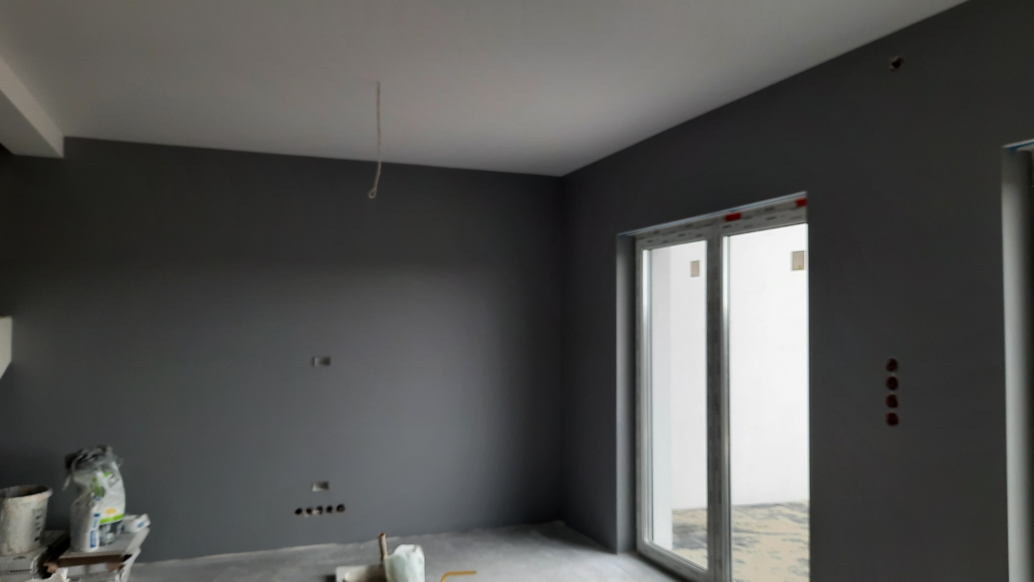 Pomieszczenie po malowaniu ścian na szaro, widoczne puszki elektryczne, okno balkonowe i materiały budowlane na podłodze.