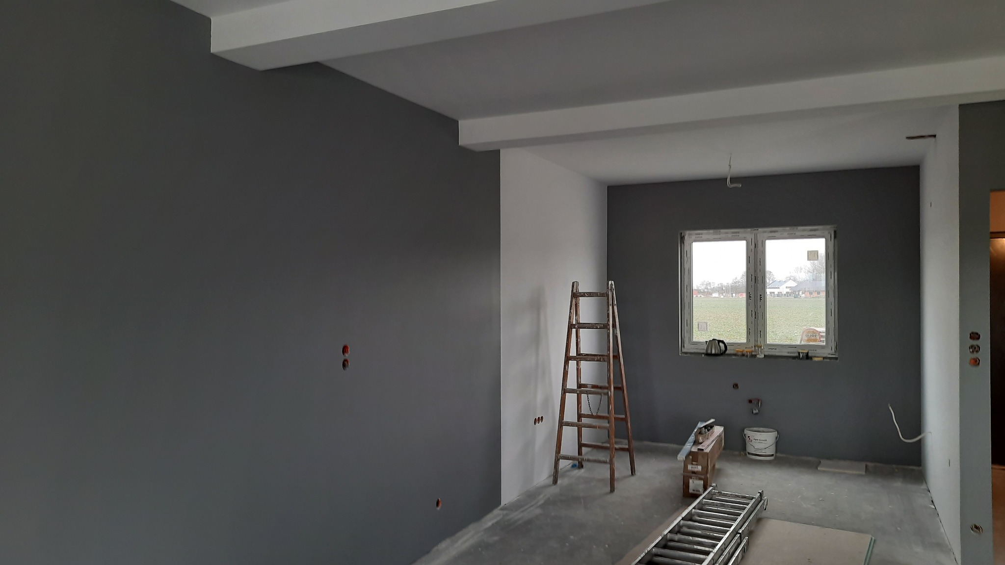 Wnętrze w Nekli po świeżym pomalowaniu ścian na szaro, widoczna drewniana drabina oparta o ścianę, częściowo zakryte okno z widokiem na zewnątrz i przygotowane narzędzia malarskie.
