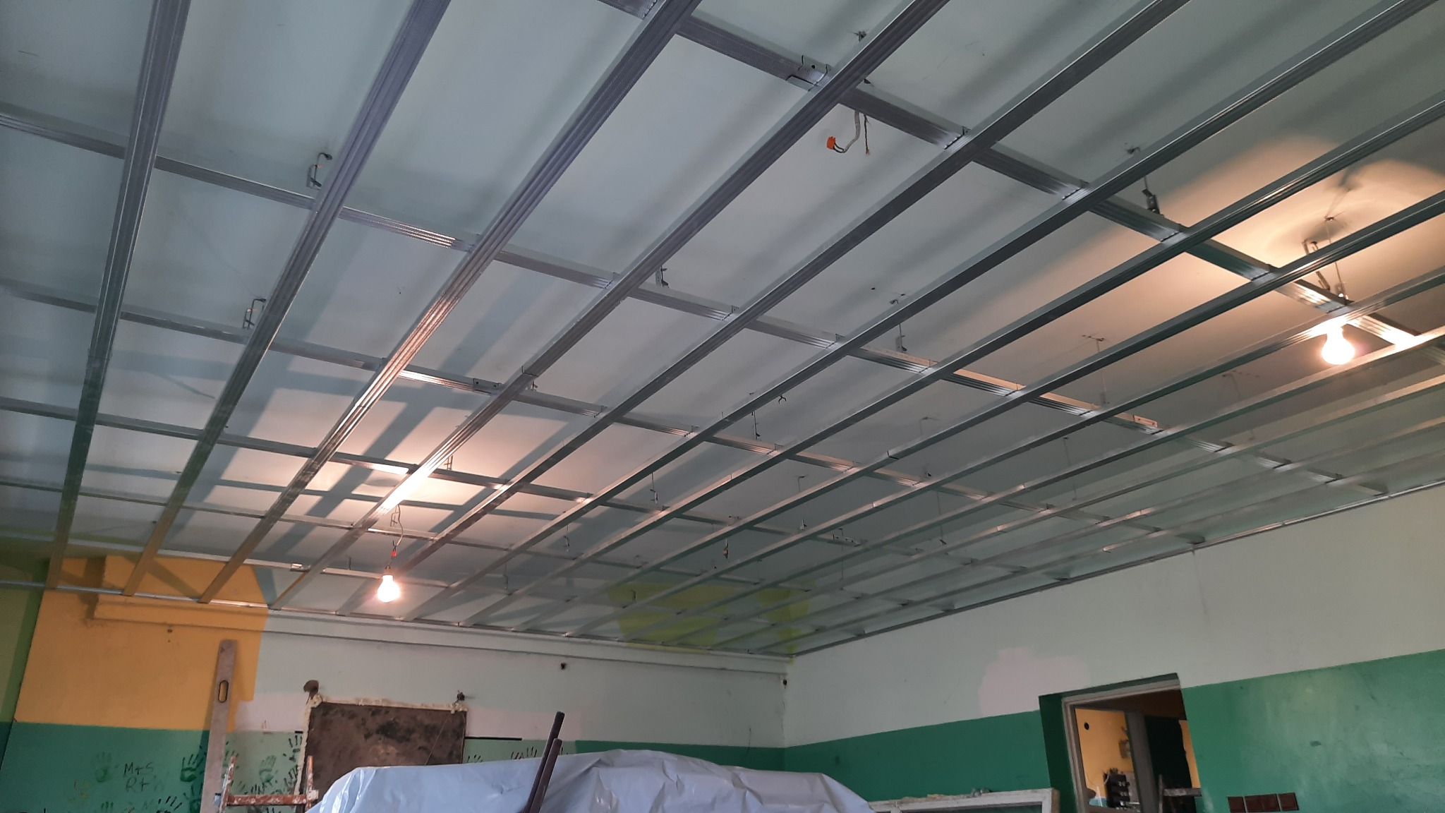 Montaż metalowej konstrukcji sufitu podwieszanego w pomieszczeniu z pomalowanymi ścianami, widoczne profile, zawiesia i oświetlenie tymczasowe.