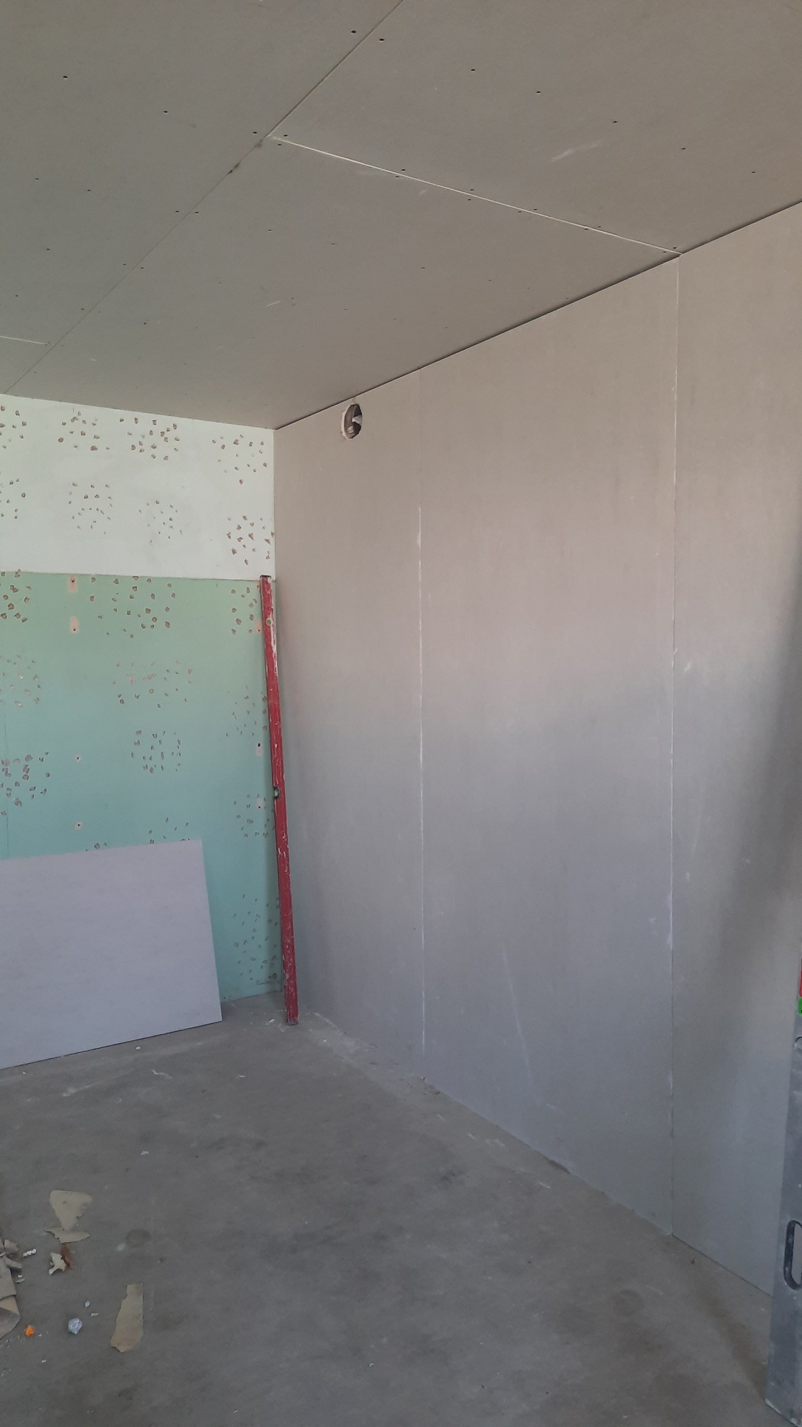 Wnętrze w trakcie prac przygotowawczych: ściany pokryte płytami gipsowo-kartonowymi, widoczny stary fragment ściany z pozostałościami tapety, poziomica oparta o ścianę, surowy beton na podłodze.