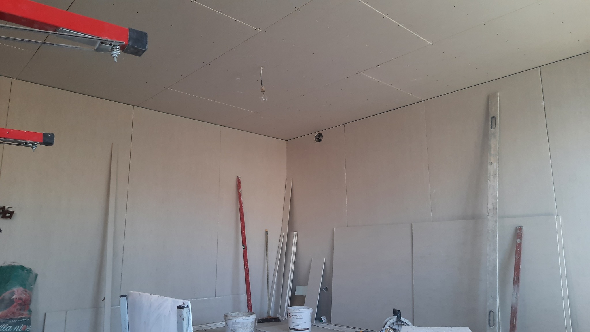 Wnętrze w trakcie montażu płyt kartonowo-gipsowych na ścianach i suficie, widoczne profile, poziomice i wiadro z zaprawą.