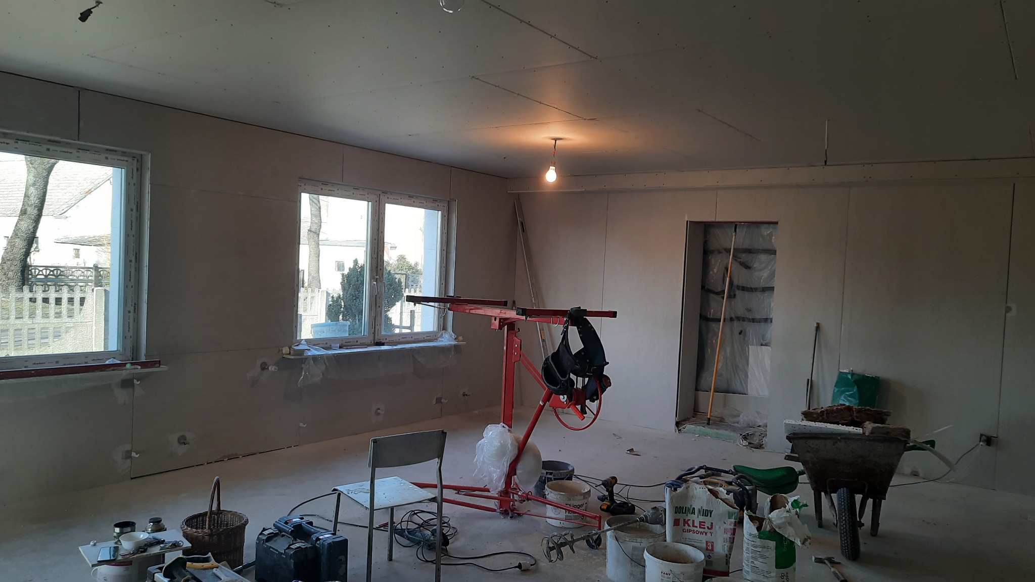 Wnętrze w trakcie prac wykończeniowych: zamontowane płyty gipsowo-kartonowe, podnośnik do płyt, wiadro z zaprawą, taczka z materiałami budowlanymi, okna z widokiem na drzewa.
