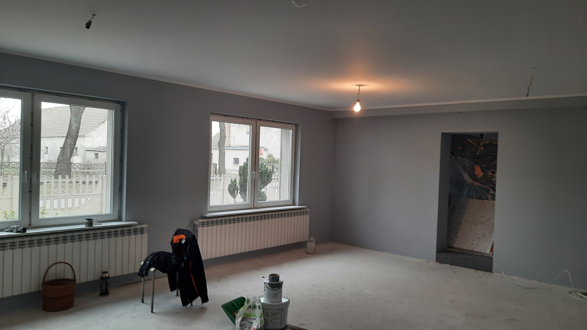 Wnętrze pomalowanego na szaro pokoju z dwoma oknami, grzejnikami, wiszącą żarówką i śladami farby na podłodze, z widocznymi akcesoriami malarskimi i odzieżą roboczą.