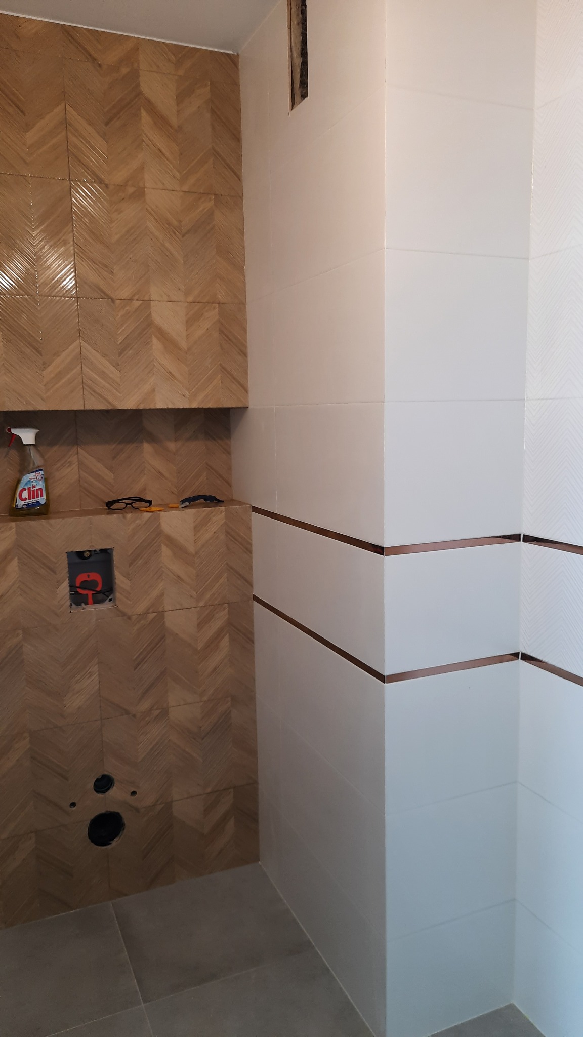 Ściana łazienki wyłożona płytkami: po lewej imitacja drewna w jodełkę, po prawej białe płytki z poziomymi, cienkimi, miedzianymi paskami. Na półce butelka płynu do czyszczenia, okulary i narzędzie.