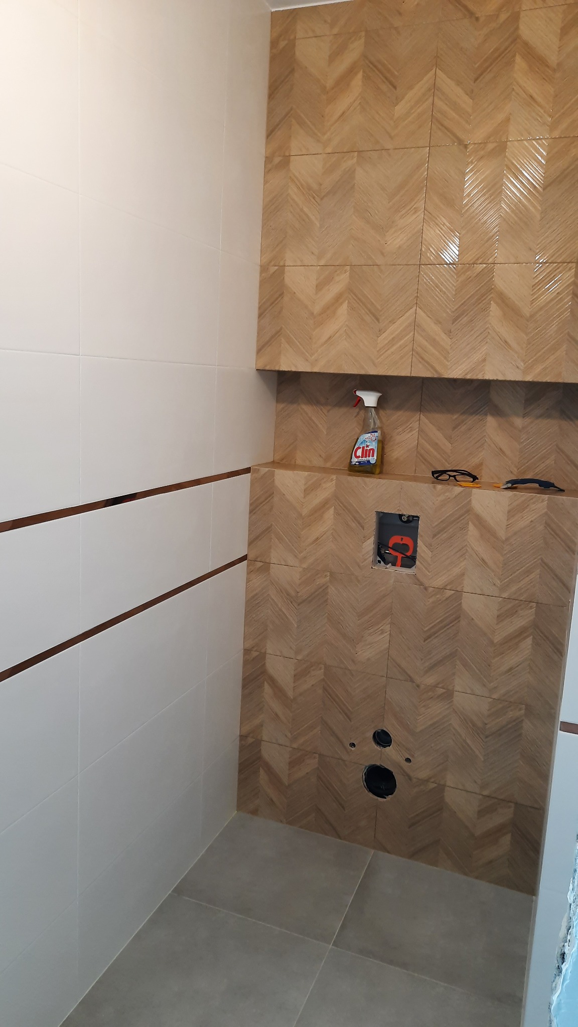 Wykończona łazienka w Nekli: białe płytki z brązowymi paskami oraz ściana z płytek imitujących drewno ułożonych we wzór jodełki. Na półce butelka płynu do czyszczenia i narzędzia glazurnicze...