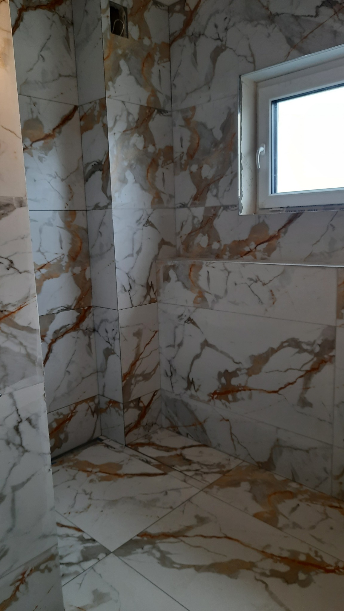 Wykończona łazienka z białymi płytkami imitującymi marmur z brązowymi żyłkami, widoczne okno z białym profilem.