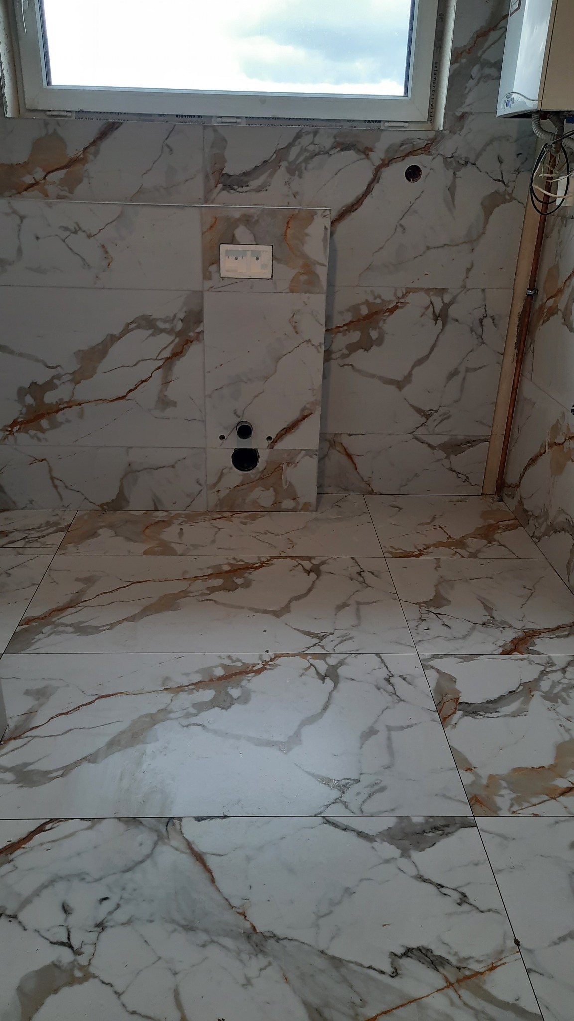 Wykończona łazienka z marmurowymi płytkami podłogowymi i ściennymi, widoczny stelaż podtynkowy WC, otwory na instalacje, jasne fugi i okno w górnej części kadru.