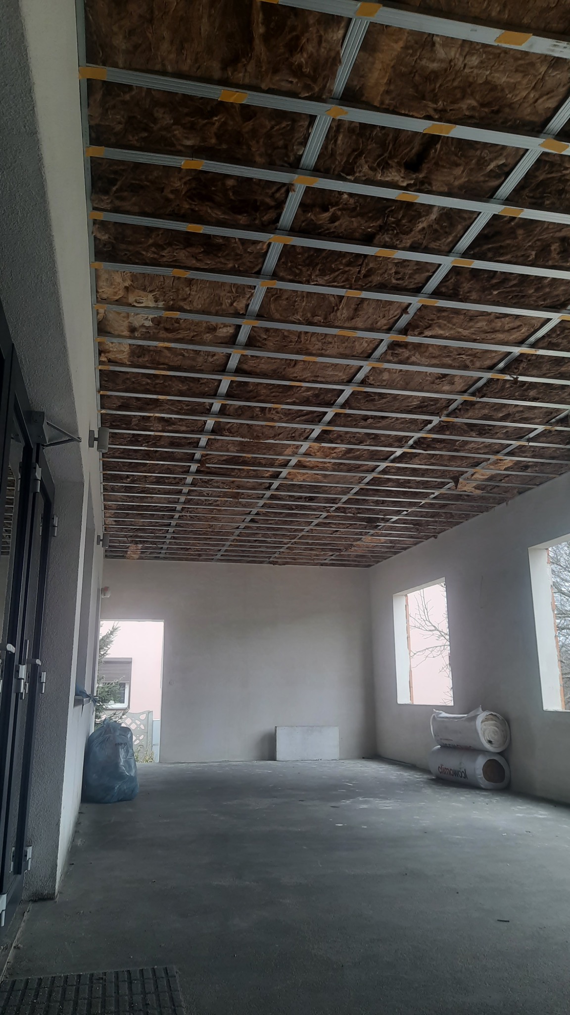 Sufit podwieszany z izolacją z wełny mineralnej w trakcie montażu, widoczne profile metalowe i fragment surowego, betonowego podłoża w pustym pomieszczeniu z oknami.