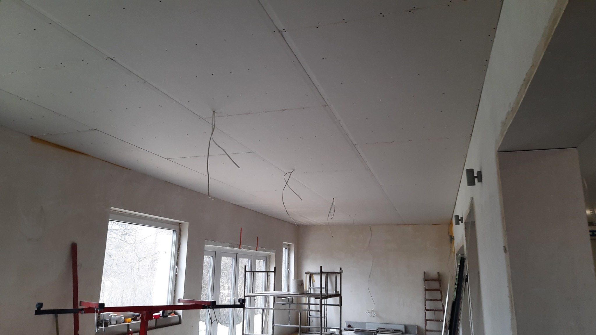 Sufit pokryty płytami gipsowo-kartonowymi w trakcie prac wykończeniowych, widoczne kable zwisające z sufitu, rusztowanie i szpachlowane ściany w pomieszczeniu z oknami.