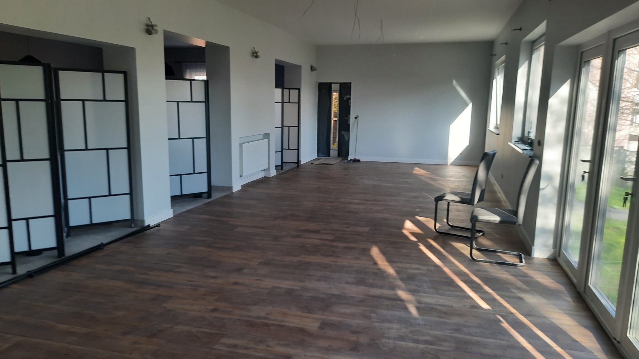 Jasne, przestronne wnętrze z panelami podłogowymi w odcieniach brązu, białymi ścianami i dużymi oknami wpuszczającymi promienie słoneczne. Widoczne dwa krzesła o nowoczesnym designie...