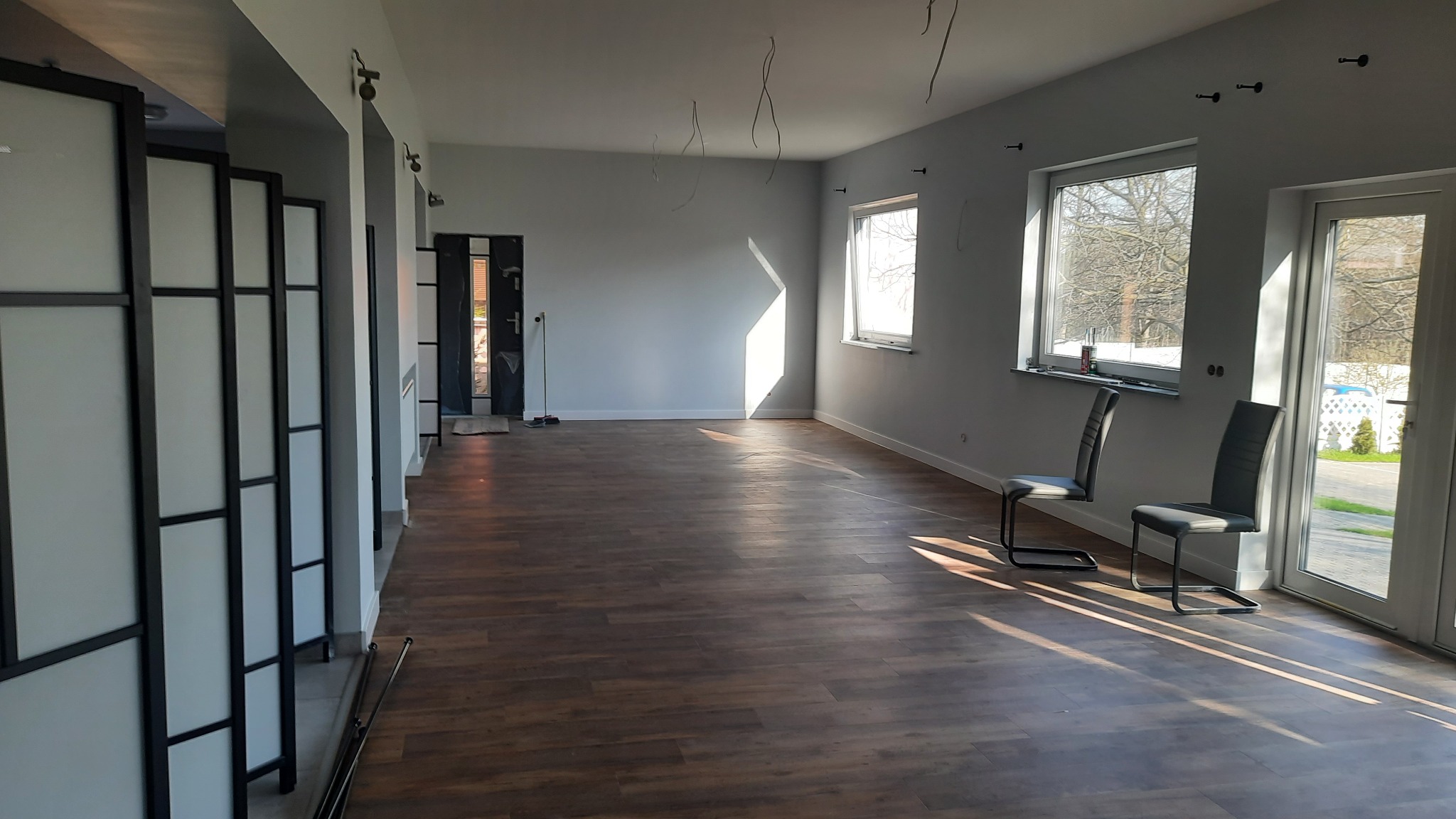 Jasne, przestronne wnętrze po remoncie z panelami podłogowymi w odcieniach brązu i szarości, z widocznymi śladami po instalacjach elektrycznych na suficie, minimalistycznymi krzesłami...