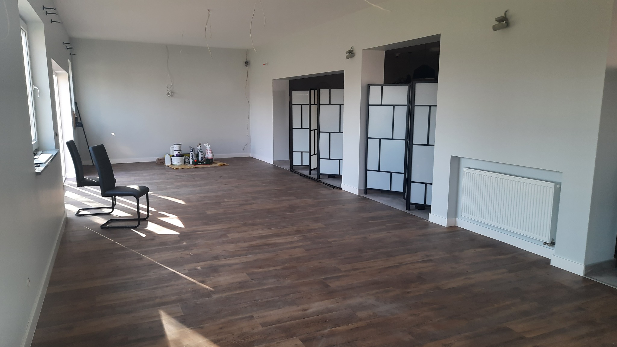 Wnętrze po malowaniu na jasny kolor z widocznymi dwoma krzesłami i pojemnikami z farbą w rogu pomieszczenia, na podłodze panele imitujące drewno.