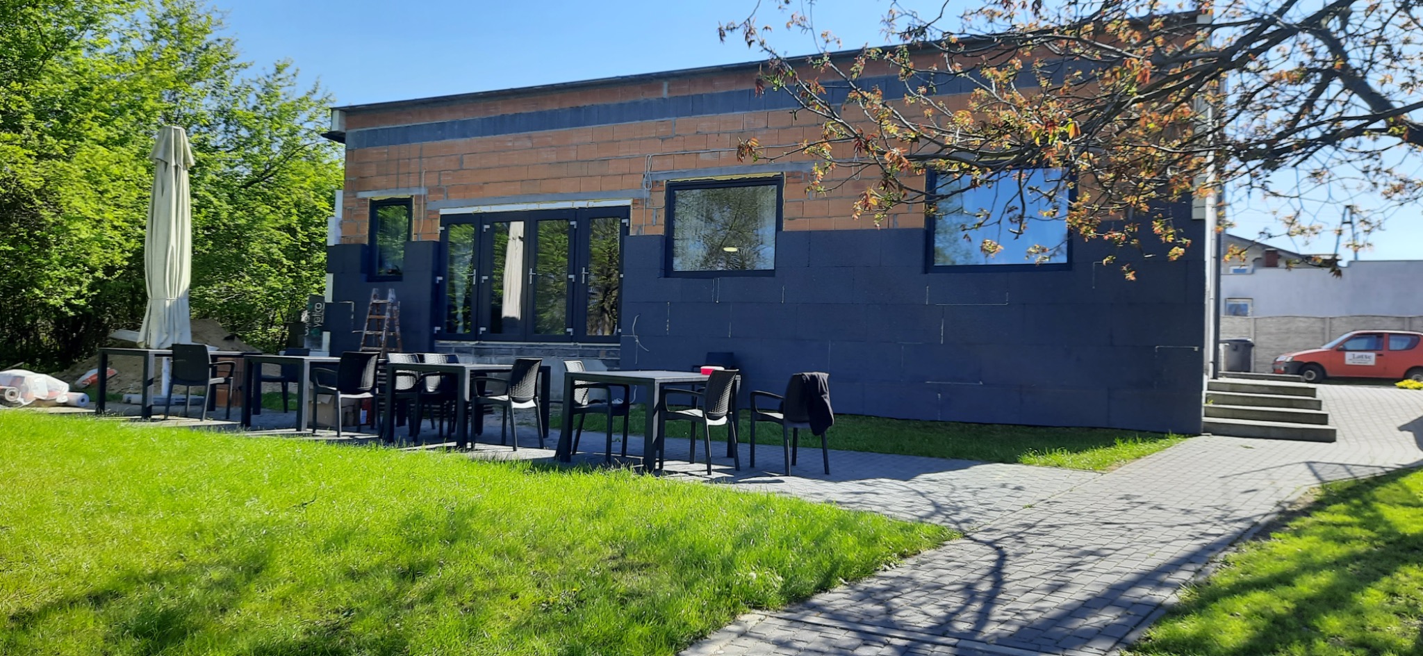 Budynek w trakcie ocieplania elewacji, widoczne płyty styropianowe i częściowo pomalowana dolna część, na zewnątrz ustawione stoliki i krzesła.