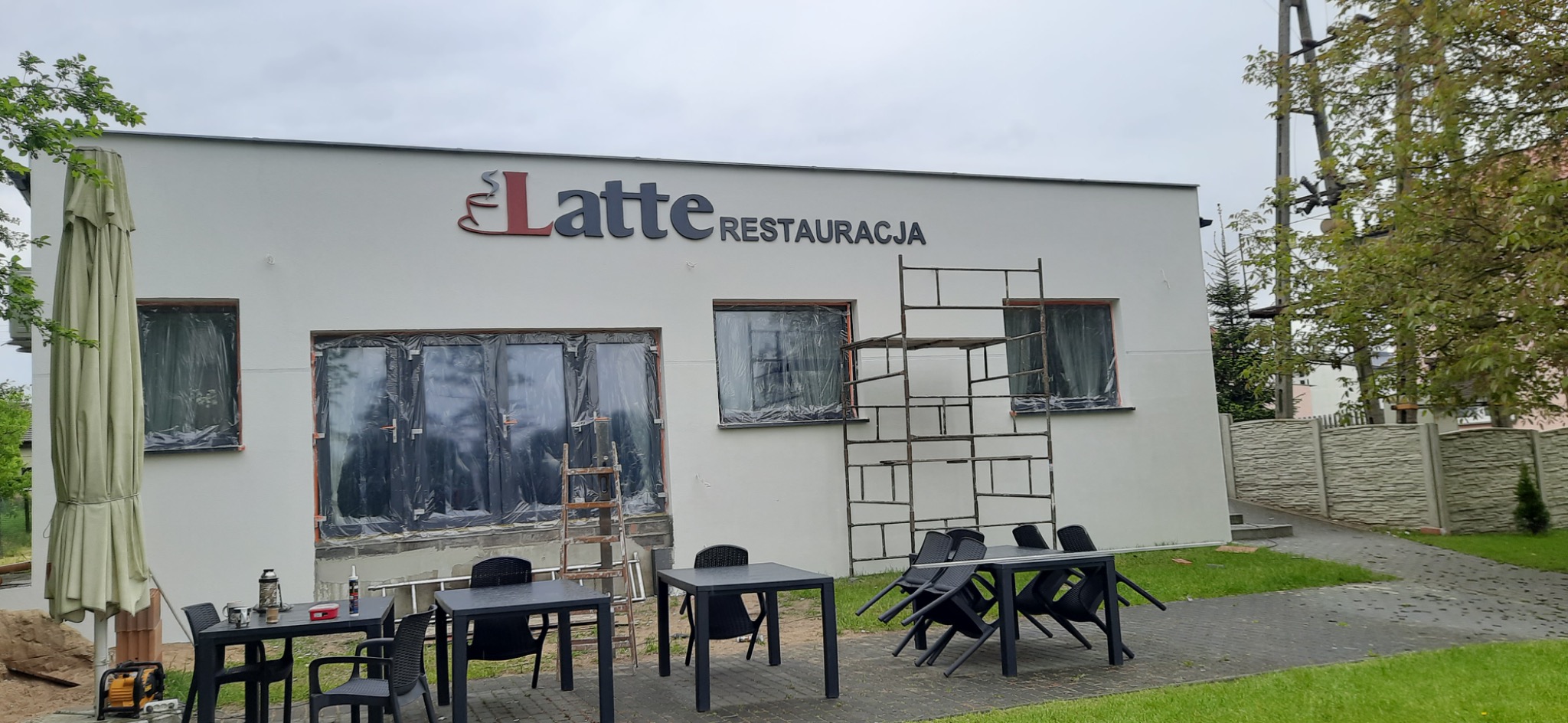 Biały budynek restauracji z zakrytymi folią oknami, rusztowaniem i stolikami na zewnątrz, sugerujący prace remontowe elewacji.