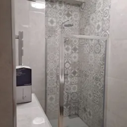 Wykończona łazienka z kabiną prysznicową, ściany wyłożone płytkami w szaro-białe wzory, obok umywalka z blatem i pudełko na szczotkę toaletową.