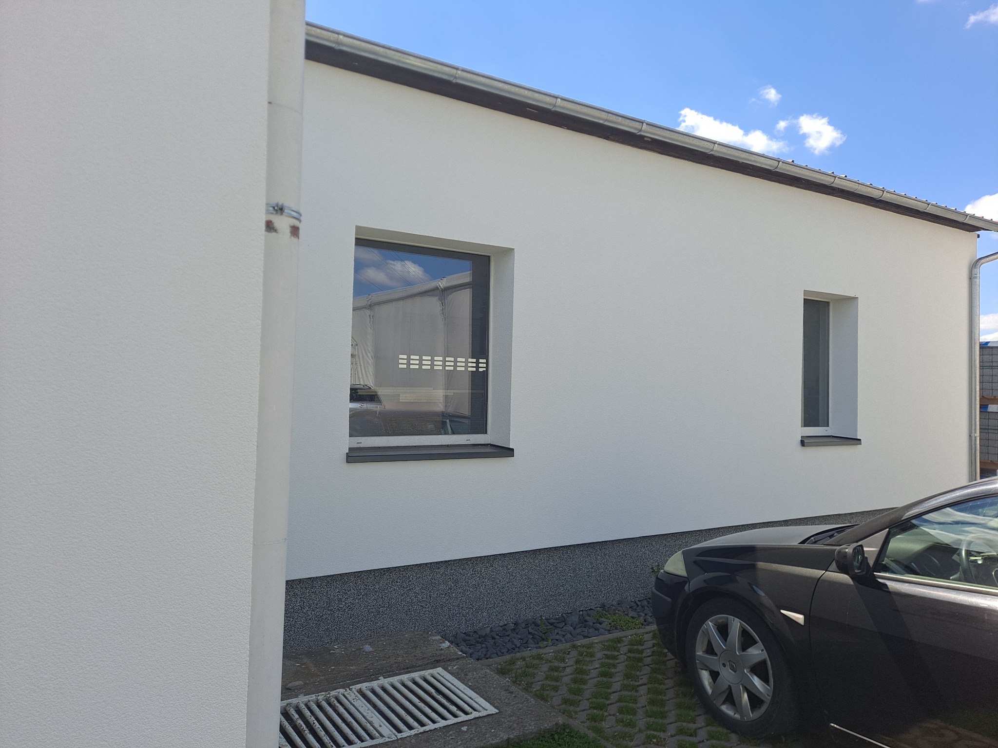 Biała elewacja budynku z dwoma oknami, widoczny fragment czarnego samochodu, szara podmurówka i grafitowa rynna, błękitne niebo z chmurami odbija się w szybie okna.