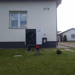 Zewnętrzna jednostka pompy ciepła Mitsubishi Zubadan zamontowana przy ścianie domu z numerem 478, z widocznymi doniczkami z kwiatami na trawniku.