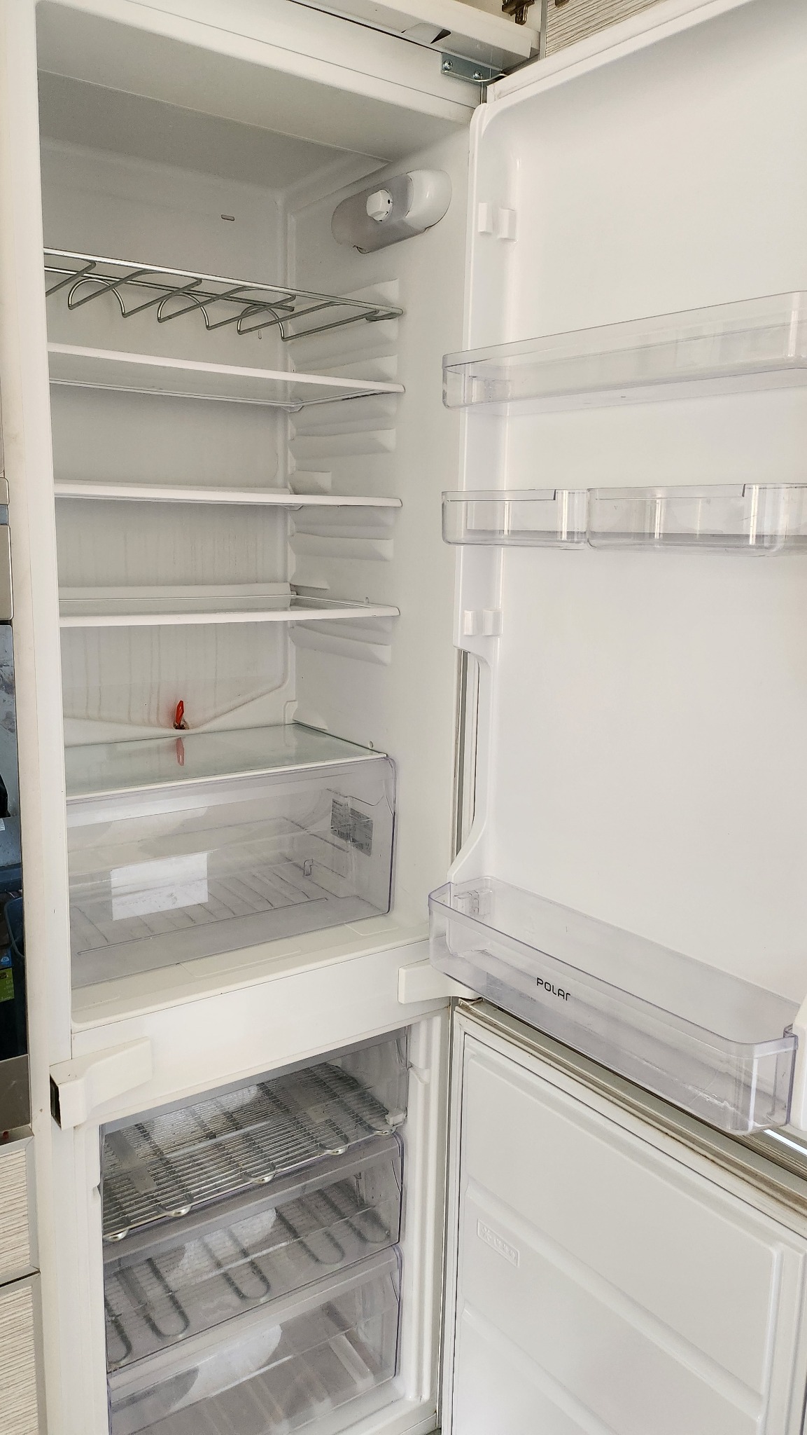 Pusta, otwarta lodówka marki Polar, widoczne szklane półki, metalowy stelaż na butelki i przezroczyste szuflady w zamrażalniku.