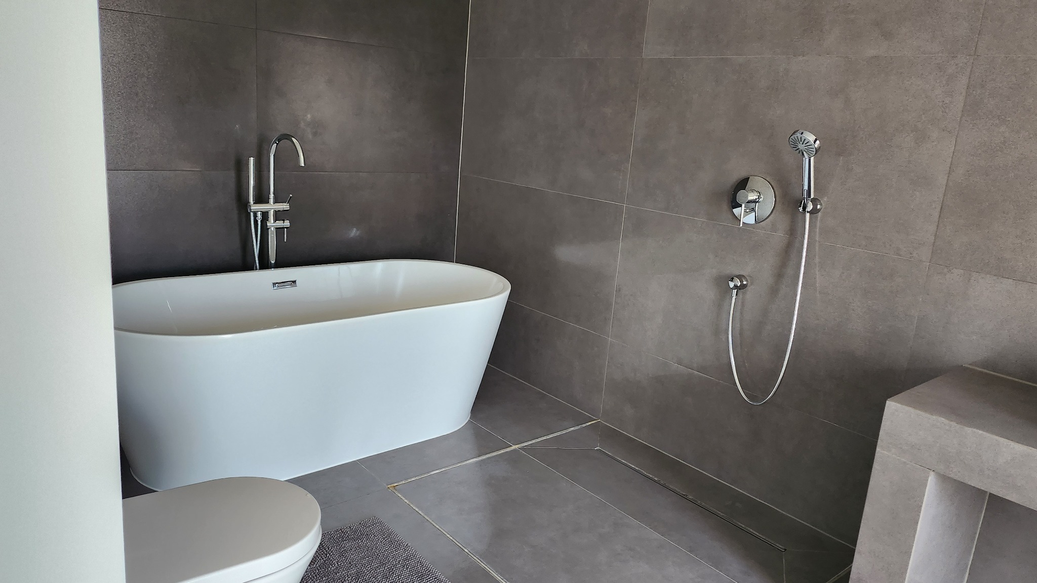 Nowoczesna łazienka z wolnostojącą białą wanną, chromowaną armaturą i szarymi płytkami podłogowymi oraz ściennymi.