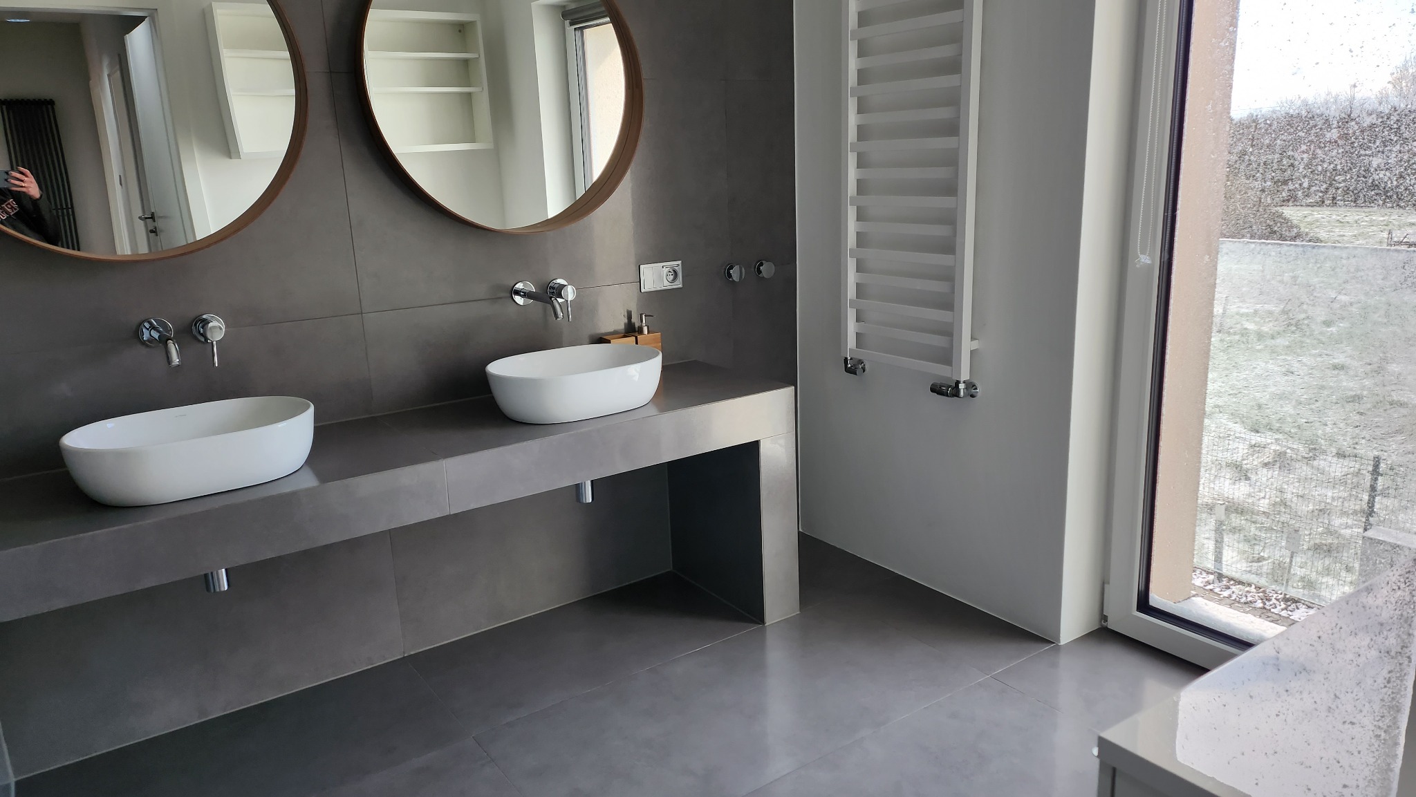 Nowoczesna łazienka z dwoma owalnymi umywalkami nablatowymi na szarym betonowym blacie, okrągłe lustra z drewnianymi ramami, grzejnik drabinkowy i okno z widokiem na zimowy krajobraz.