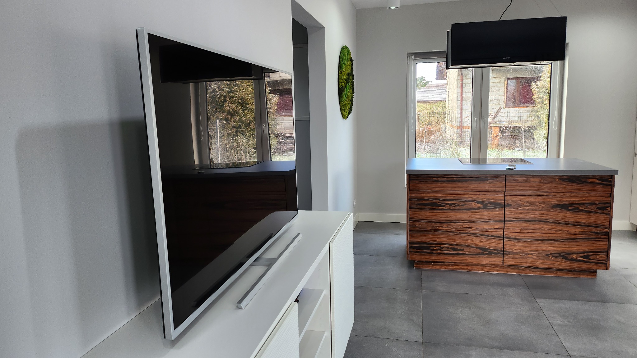 Nowoczesna kuchnia z wyspą i telewizorem, widok przez okno na zewnątrz, minimalistyczny wystrój z mchem na ścianie.