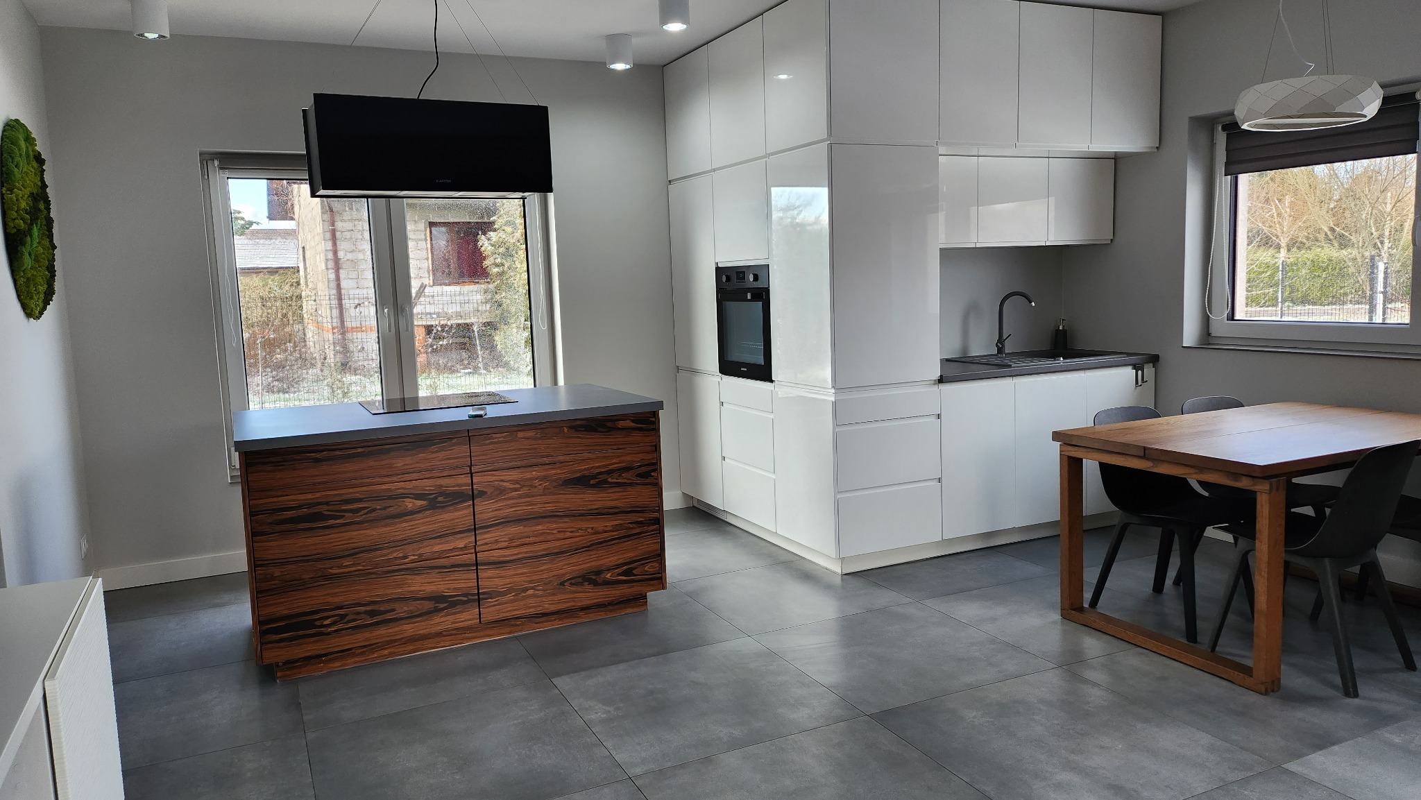 Nowoczesna kuchnia z wyspą z forniru w kolorze ciemnego drewna, białe szafki, czarny piekarnik i stół z krzesłami na szarych płytkach.