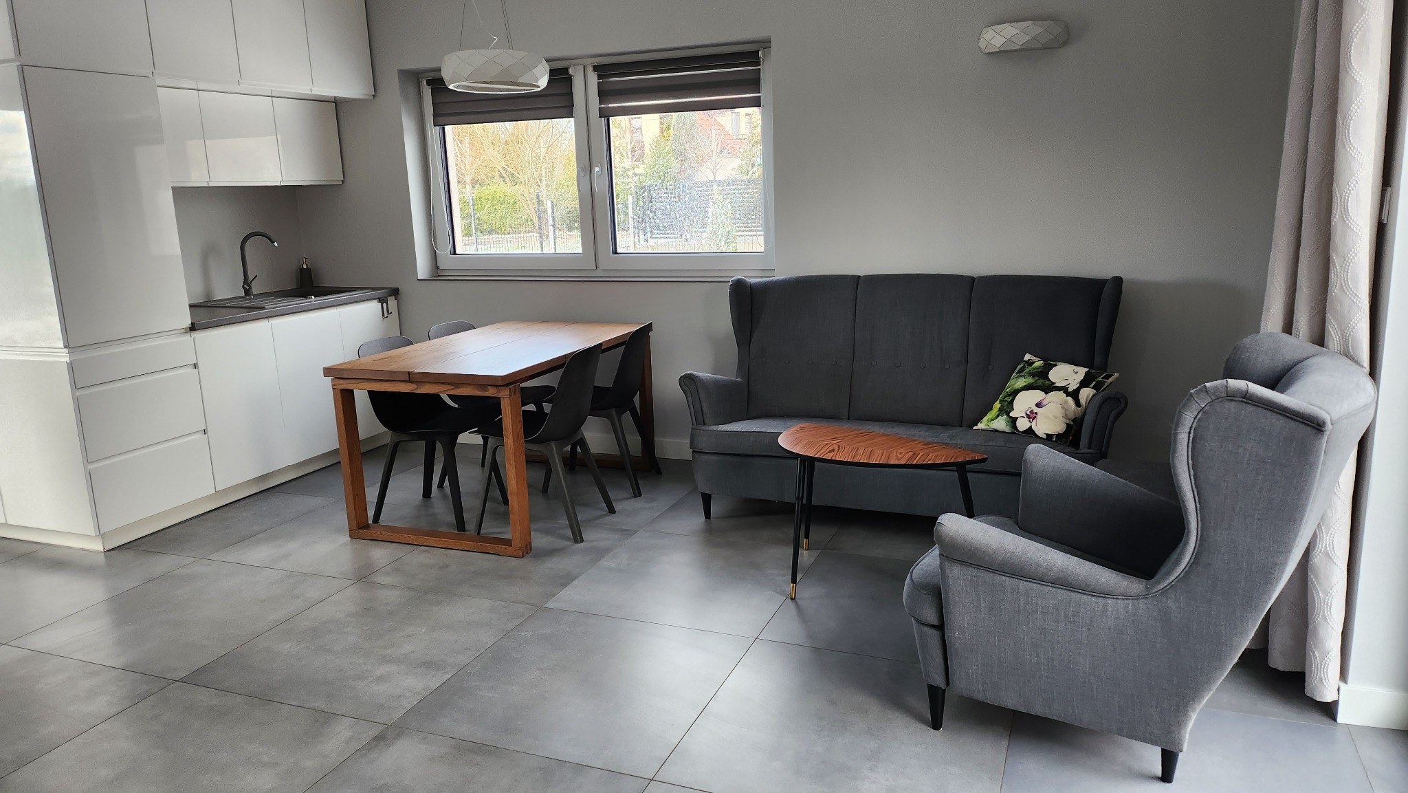 Jasne wnętrze mieszkania z szarymi meblami, drewnianym stołem i białymi szafkami kuchennymi, widok z okna na ogród.