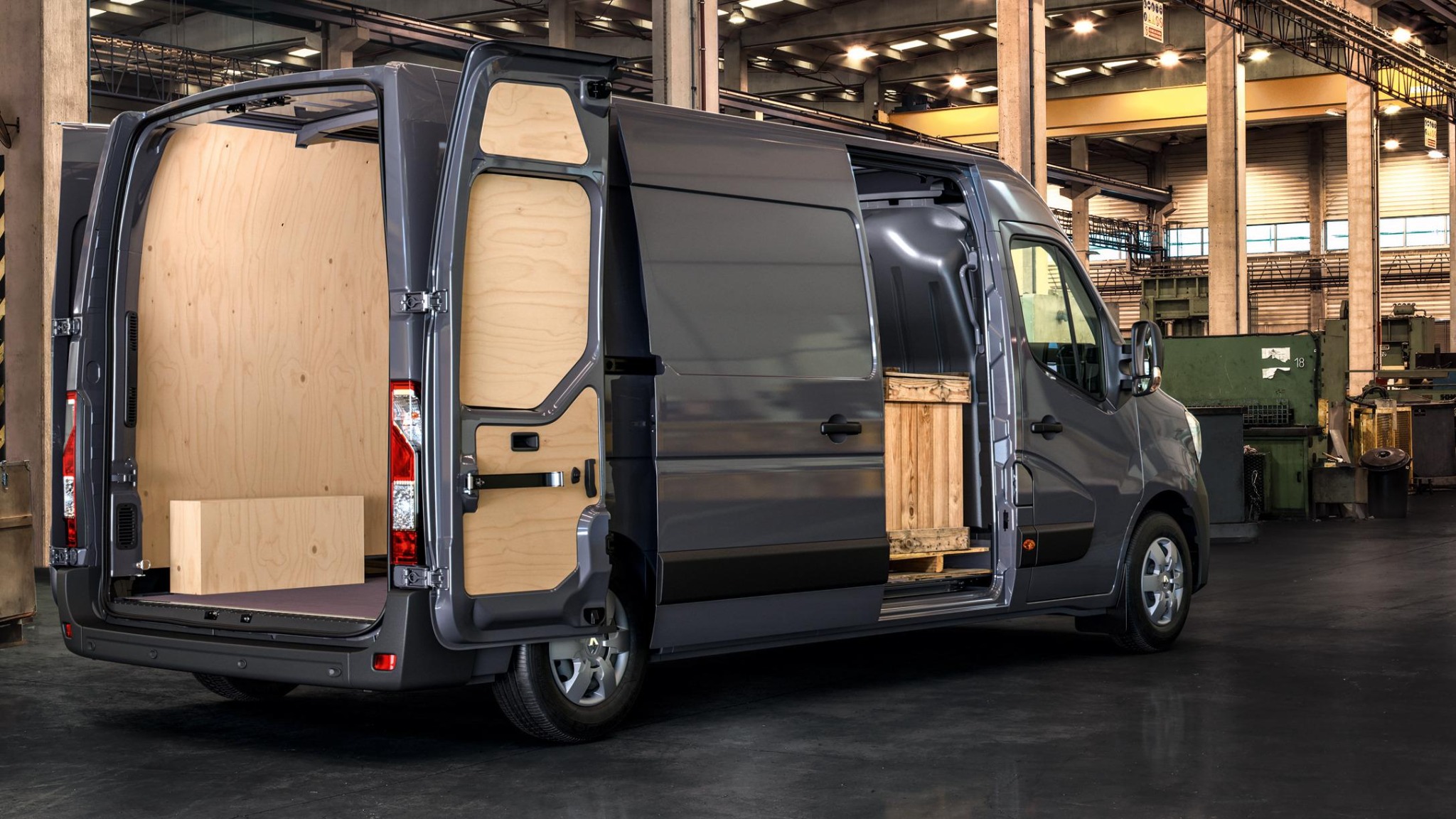 Szary van z otwartymi tylnymi i bocznymi drzwiami, widoczne drewniane obicie wnętrza i paleta z drewnianą skrzynią w środku, na tle hali przemysłowej.
