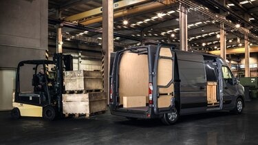 Wnętrze otwartego, szarego vana z drewnianą zabudową, obok wózek widłowy z paletami w hali magazynowej.