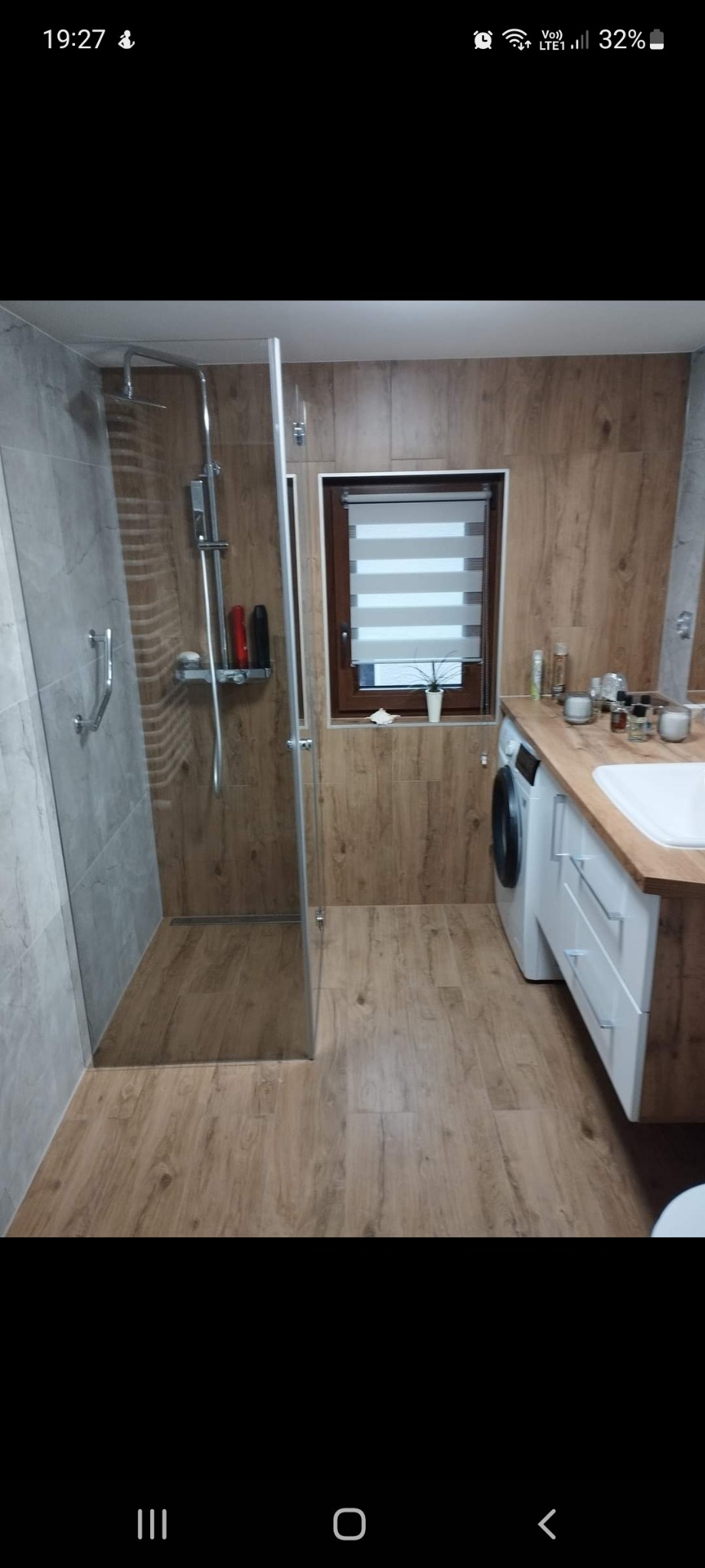 Nowoczesna łazienka z kabiną prysznicową typu walk-in, umywalką nablatową na drewnianym blacie, pralką i oknem z roletami dzień-noc.