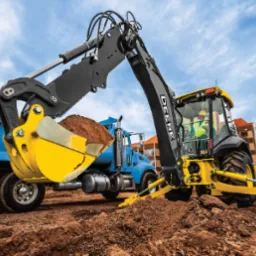 Żółto-czarna koparko-ładowarka John Deere z pełną łyżką ziemi, obok niebieska ciężarówka wywrotka, w tle budynki mieszkalne.