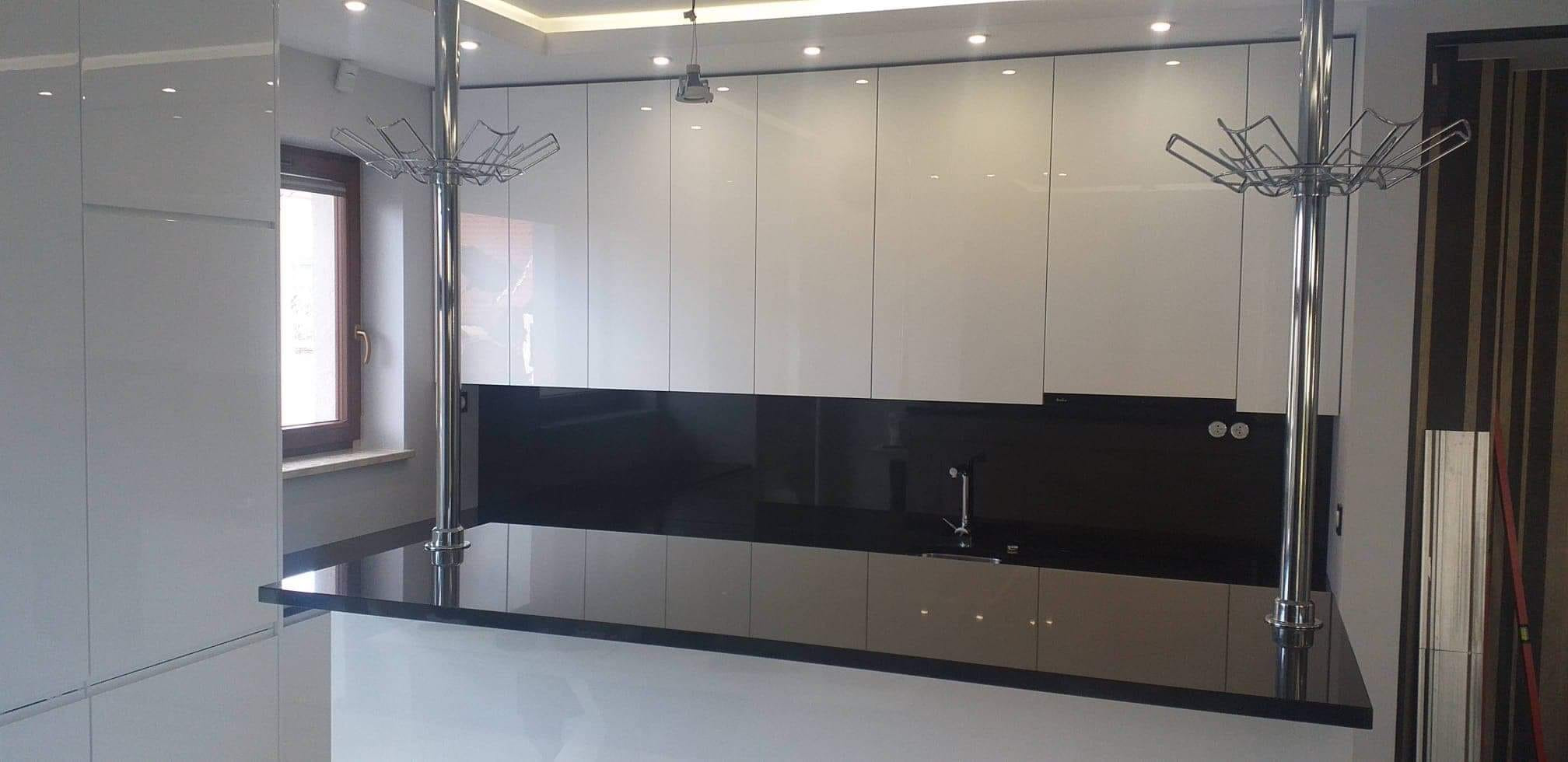 Nowoczesna kuchnia z białymi, lakierowanymi szafkami górnymi i czarnym blatem, podpartym chromowanymi słupkami z wieszakami, oświetlona punktowymi lampami sufitowymi.