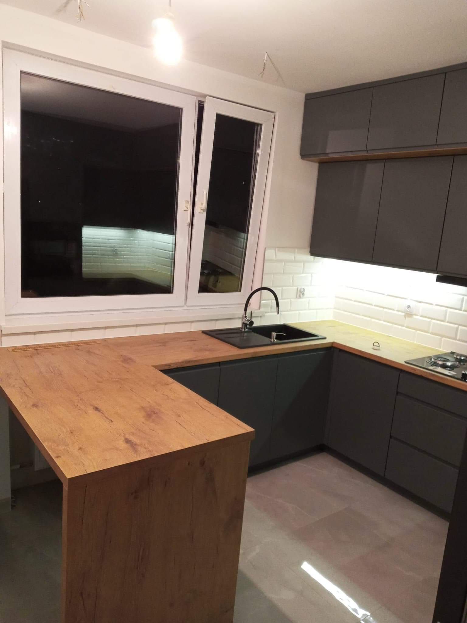 Nowoczesna kuchnia w odcieniach szarości z drewnianym blatem, białymi płytkami metro i czarną armaturą, oświetlona punktowo pod szafkami.