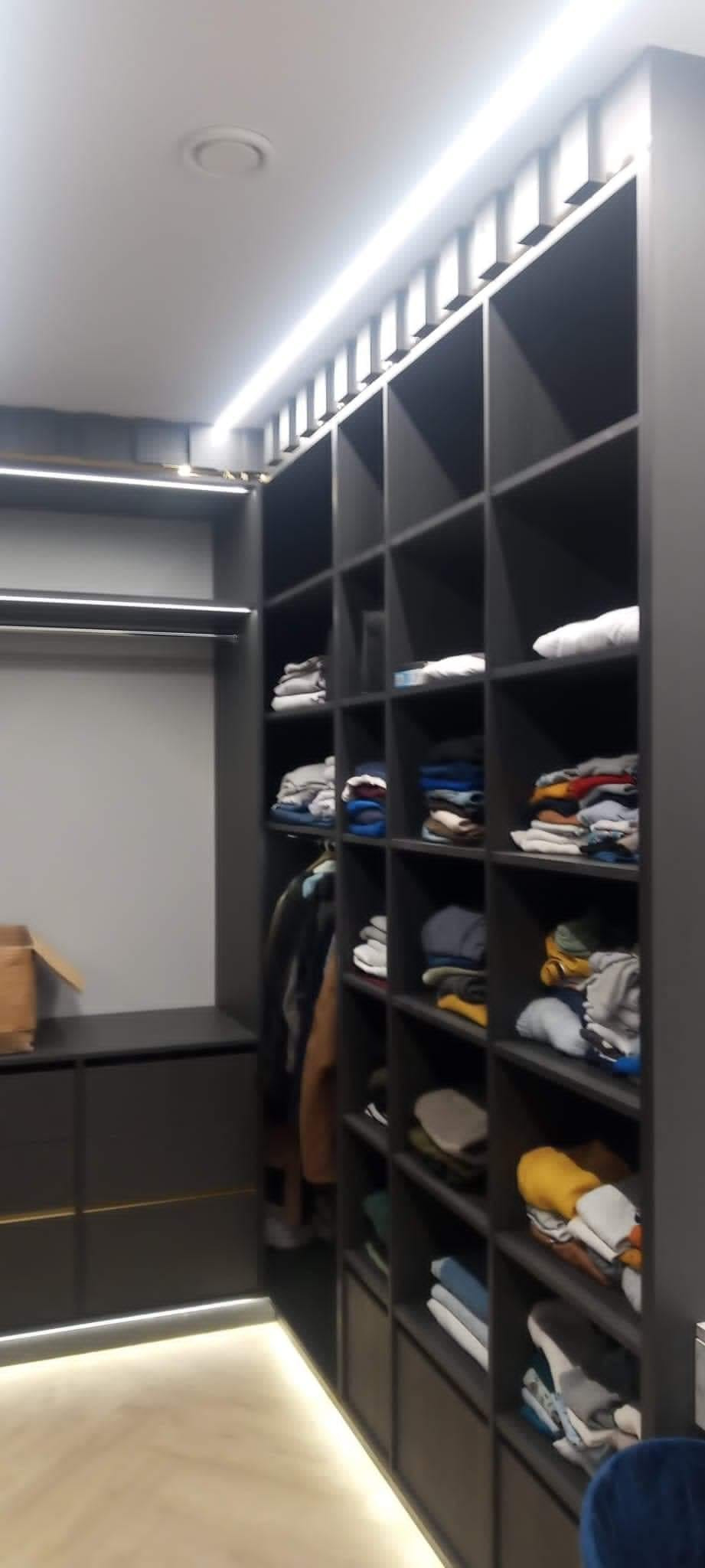 Szara, zabudowana garderoba z oświetleniem LED, półki wypełnione ubraniami i akcesoriami, widoczny fragment podłogi.