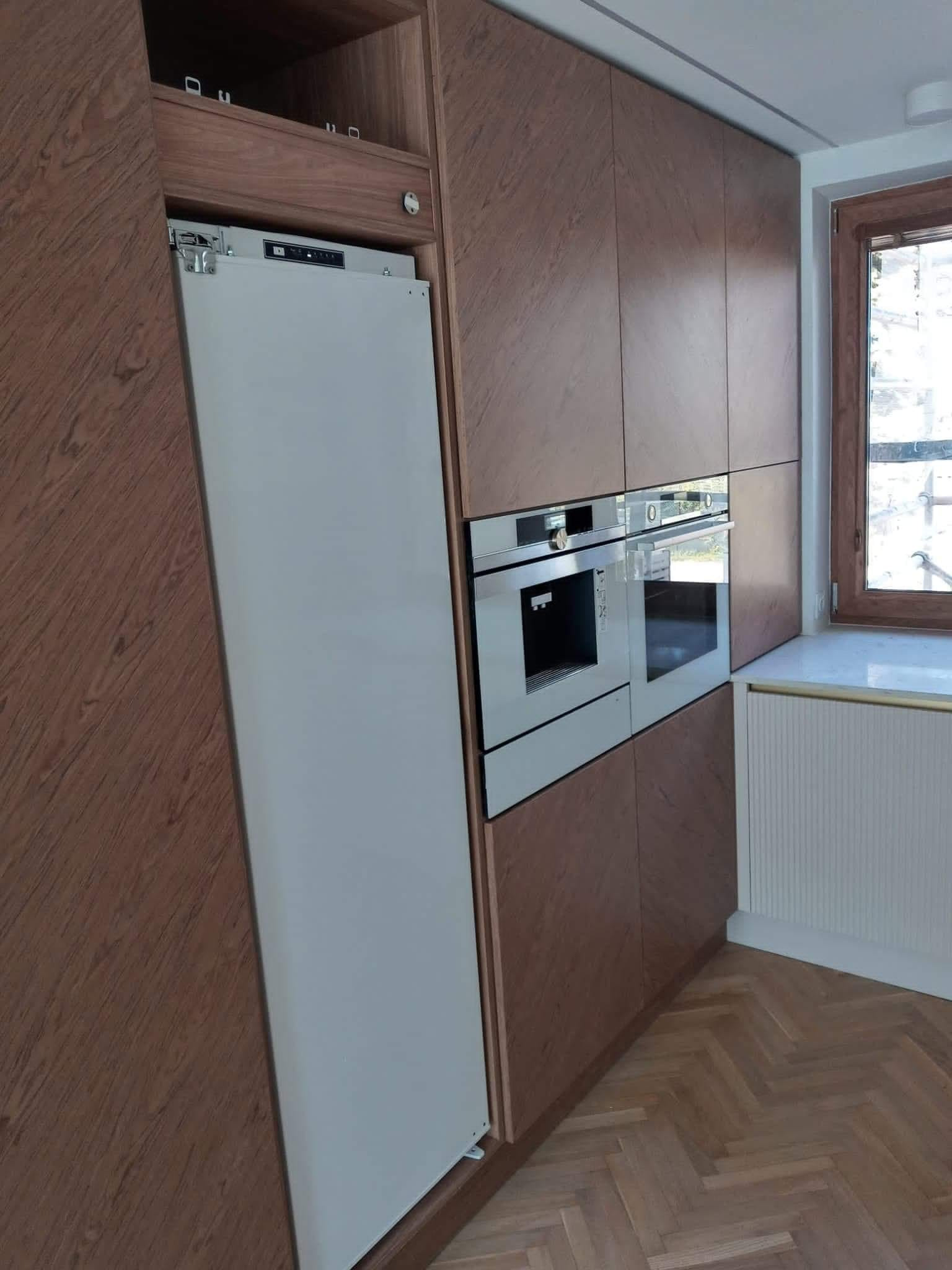 Nowoczesna kuchnia z drewnianymi szafkami, wbudowanym piekarnikiem i lodówką ukrytą za frontem szafki, parkiet w jodełkę na podłodze.