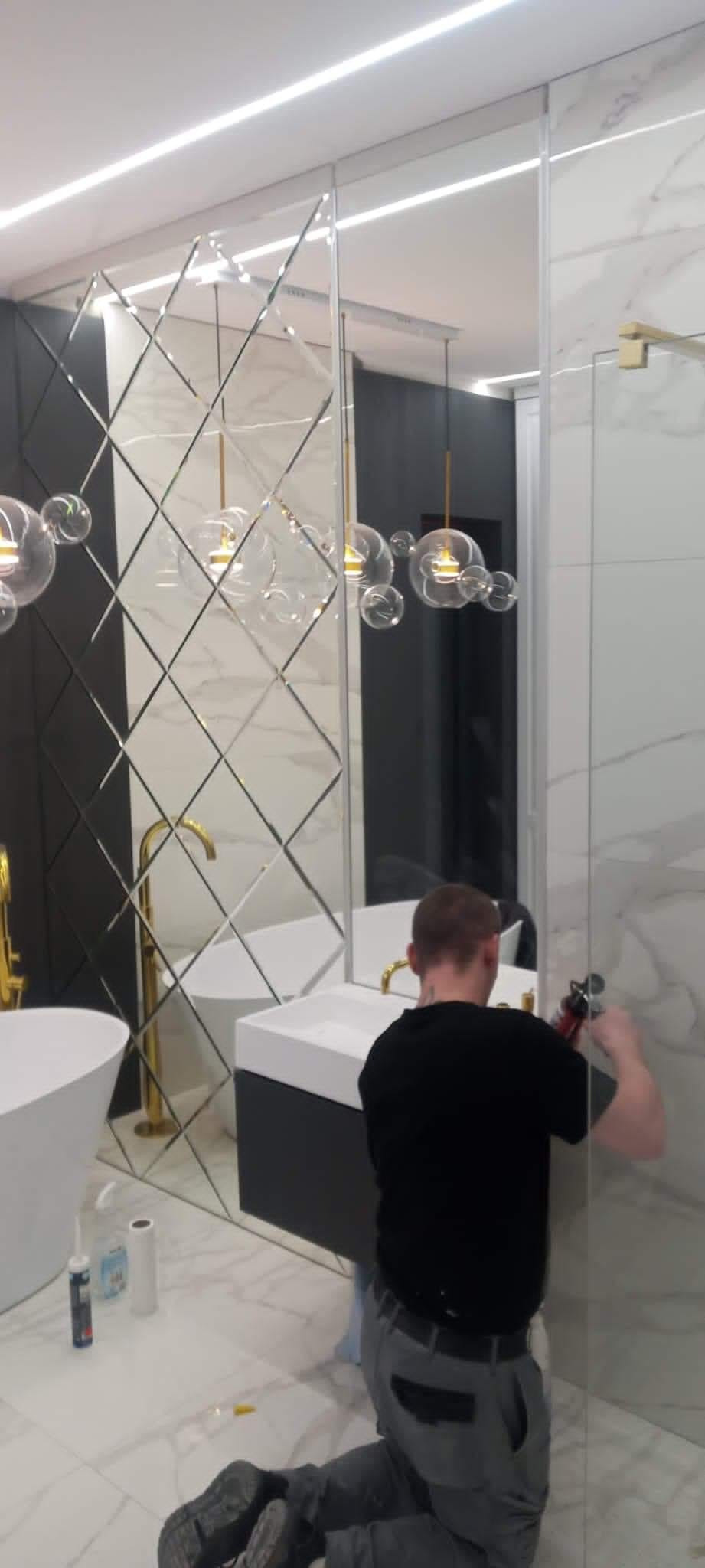 Montaż szklanej kabiny prysznicowej w nowoczesnej łazience z lustrzaną ścianą w romby, złotą armaturą i wiszącymi lampami typu bubble.